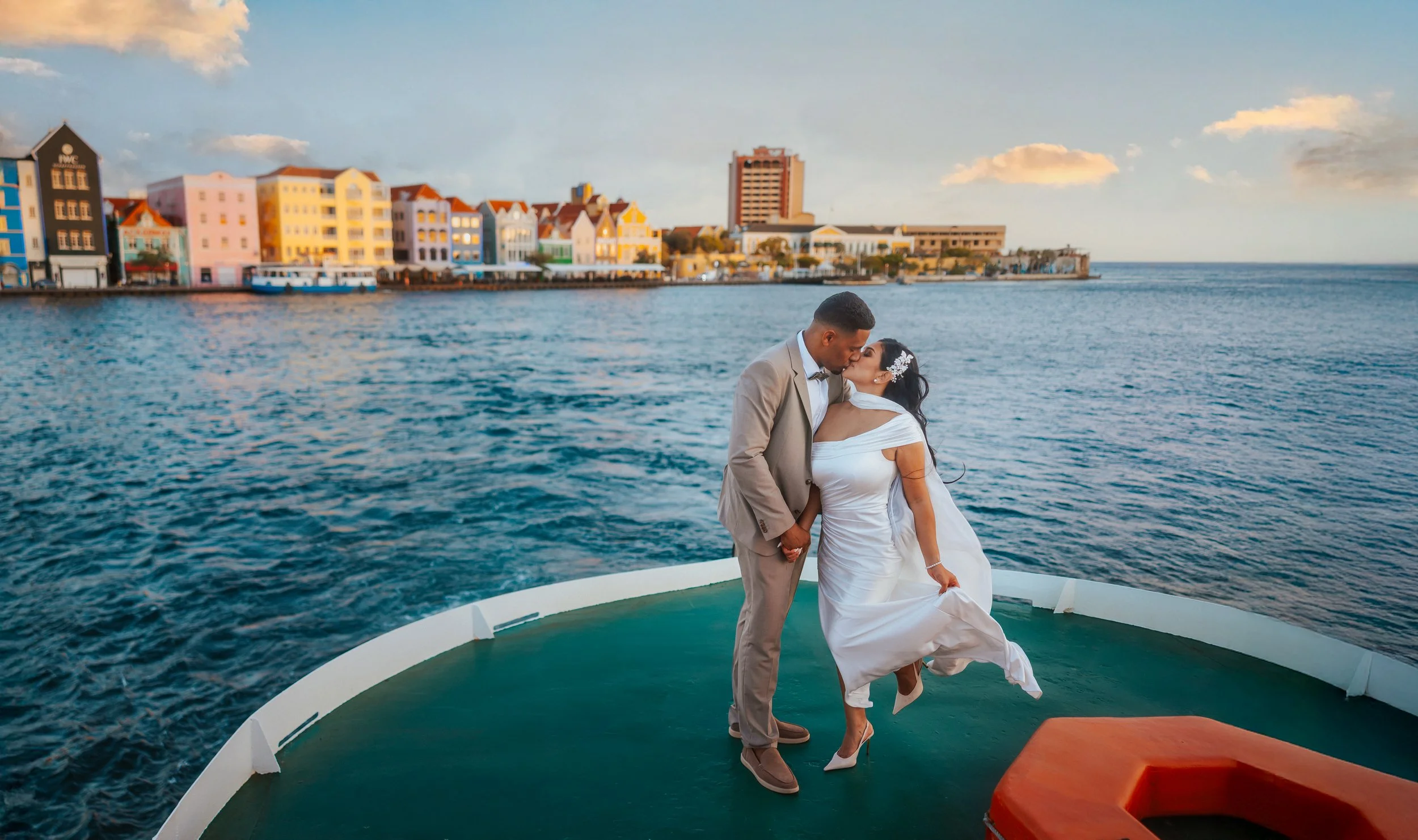 A couple dressed in wedding attire sharing a kiss on a boat with a colorful seaside town in the background.