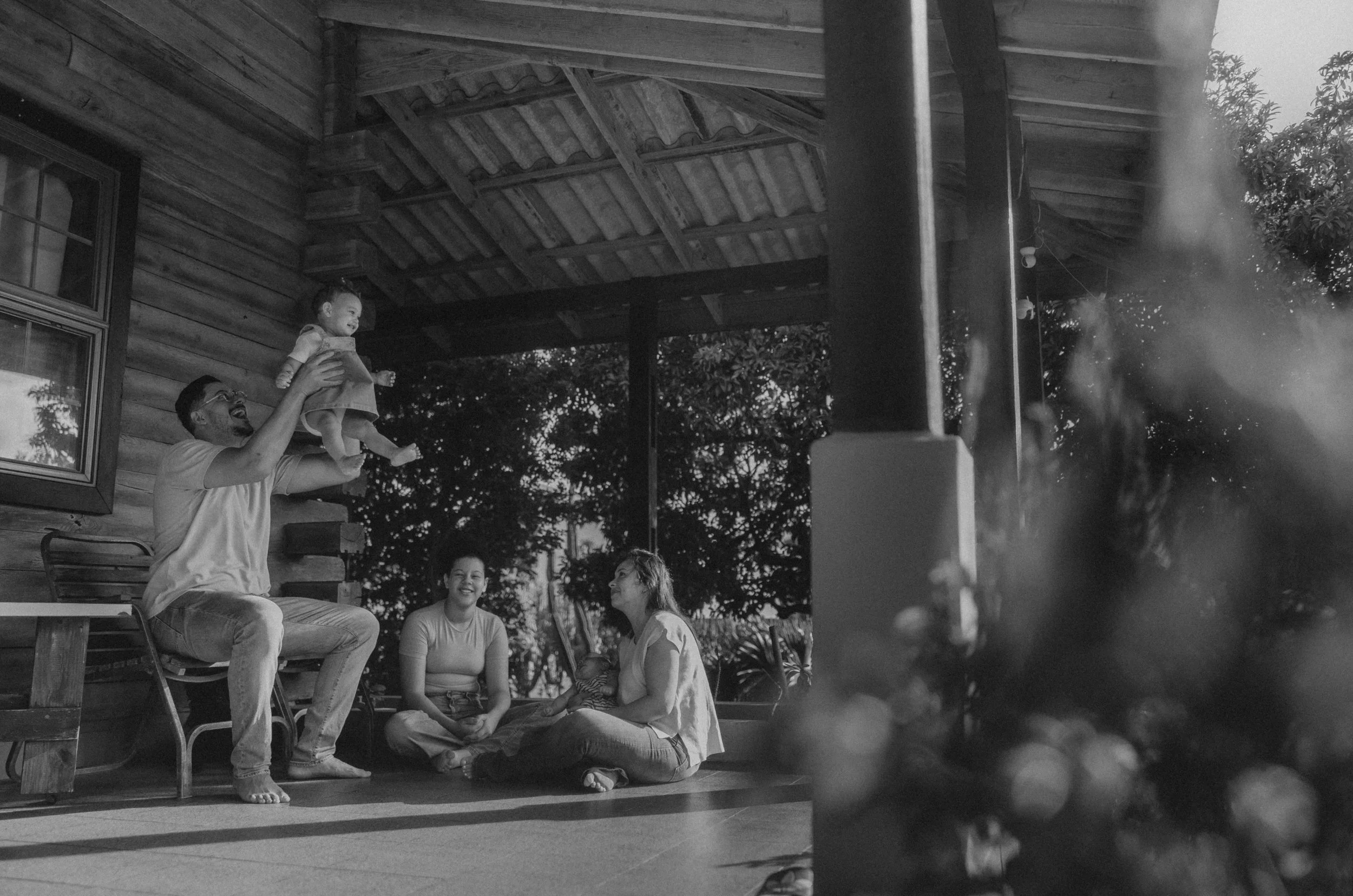 A man holding a smiling child above his head outdoors on a porch, with two women sitting on the floor nearby. The scene is in black and white, and the porch has a wooden ceiling and a window.