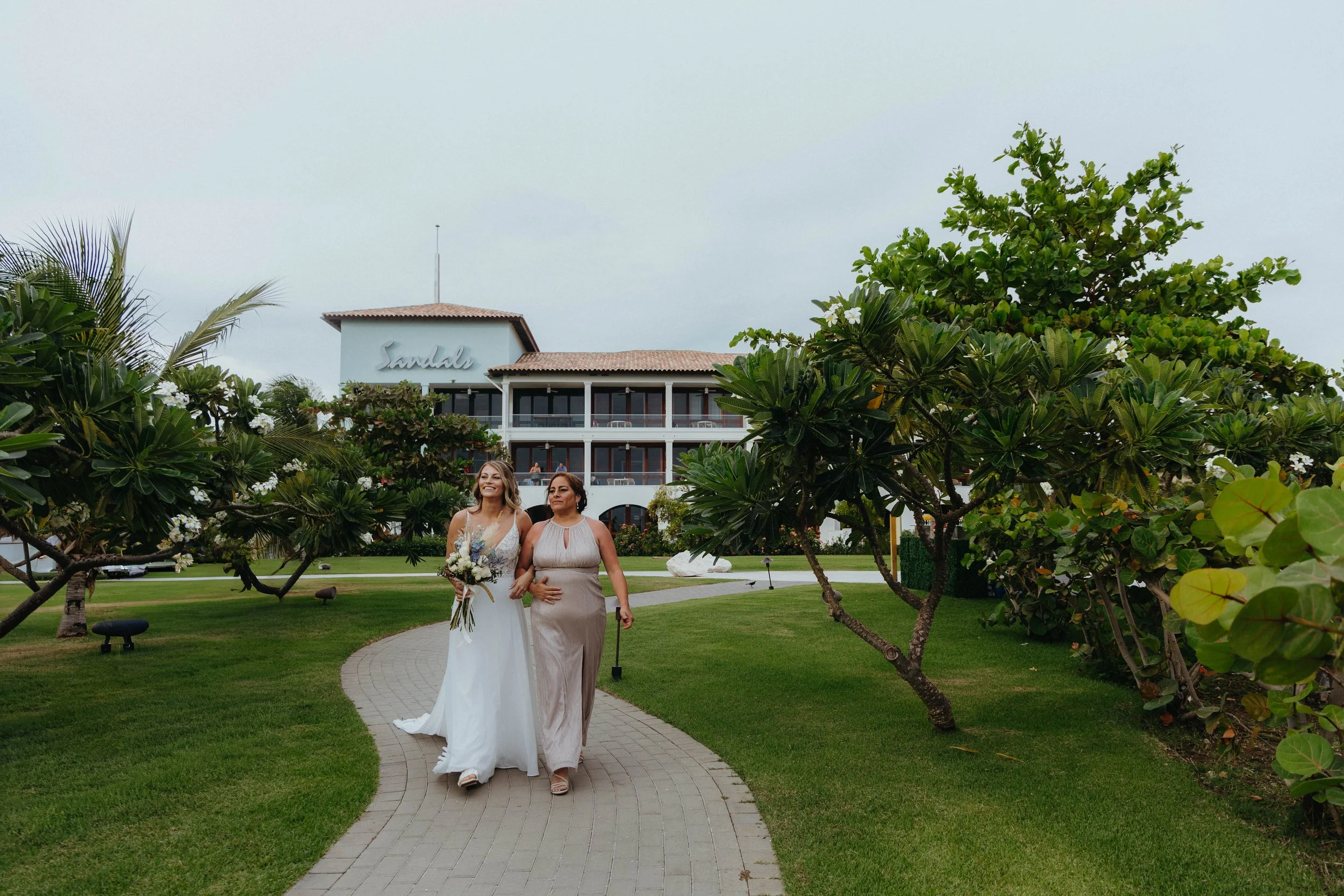 Two women walking arm in arm on a paved path in a garden, with a hotel building named 'Sandals' in the background. One woman is dressed in a white wedding gown, holding a bouquet of flowers, and the other in a light pink dress. The scene appears to b