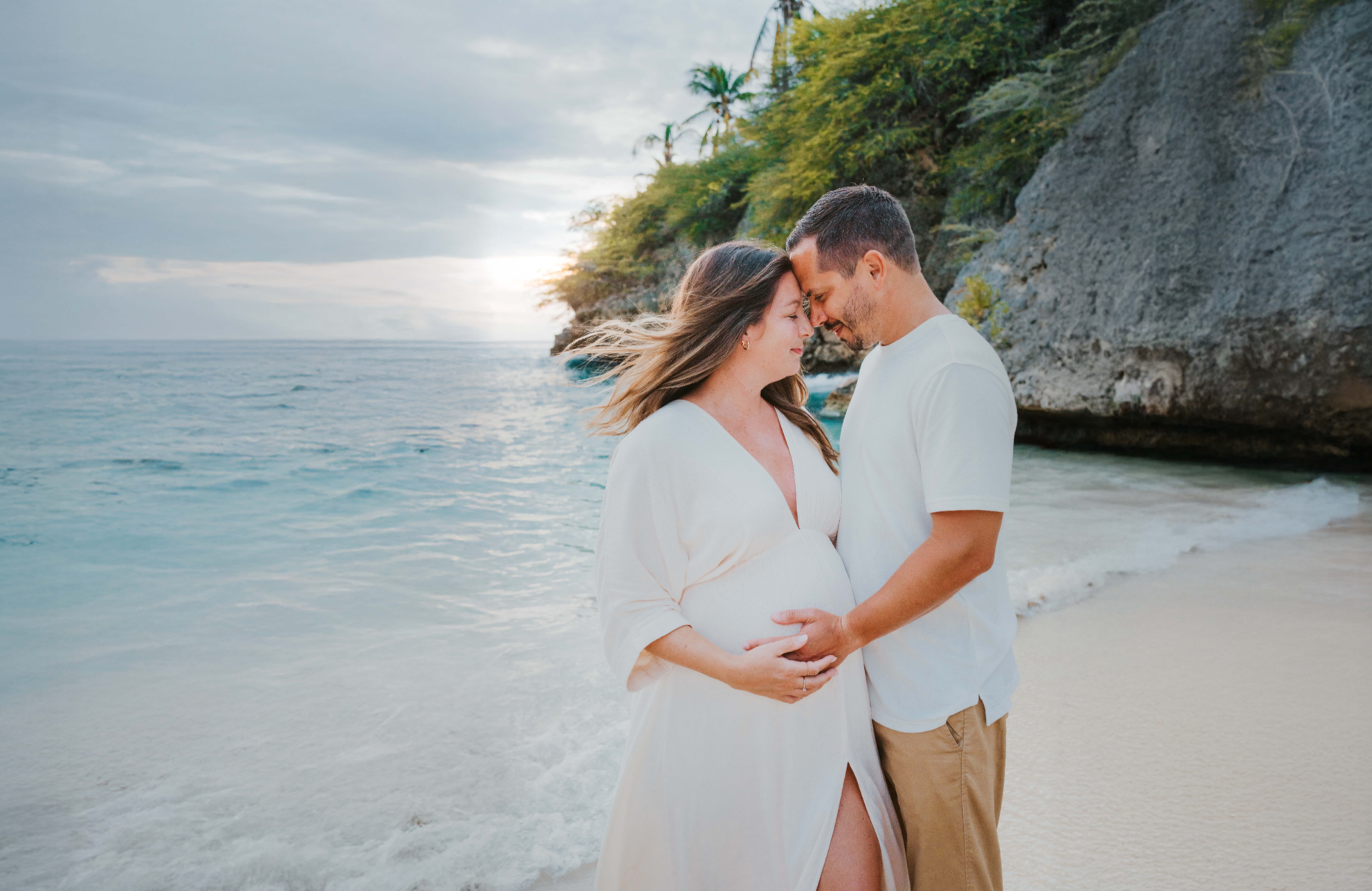 A pregnant woman and a man standing close together on the beach with their foreheads touching, holding her baby bump, near the water with cliffs and tropical trees in the background.