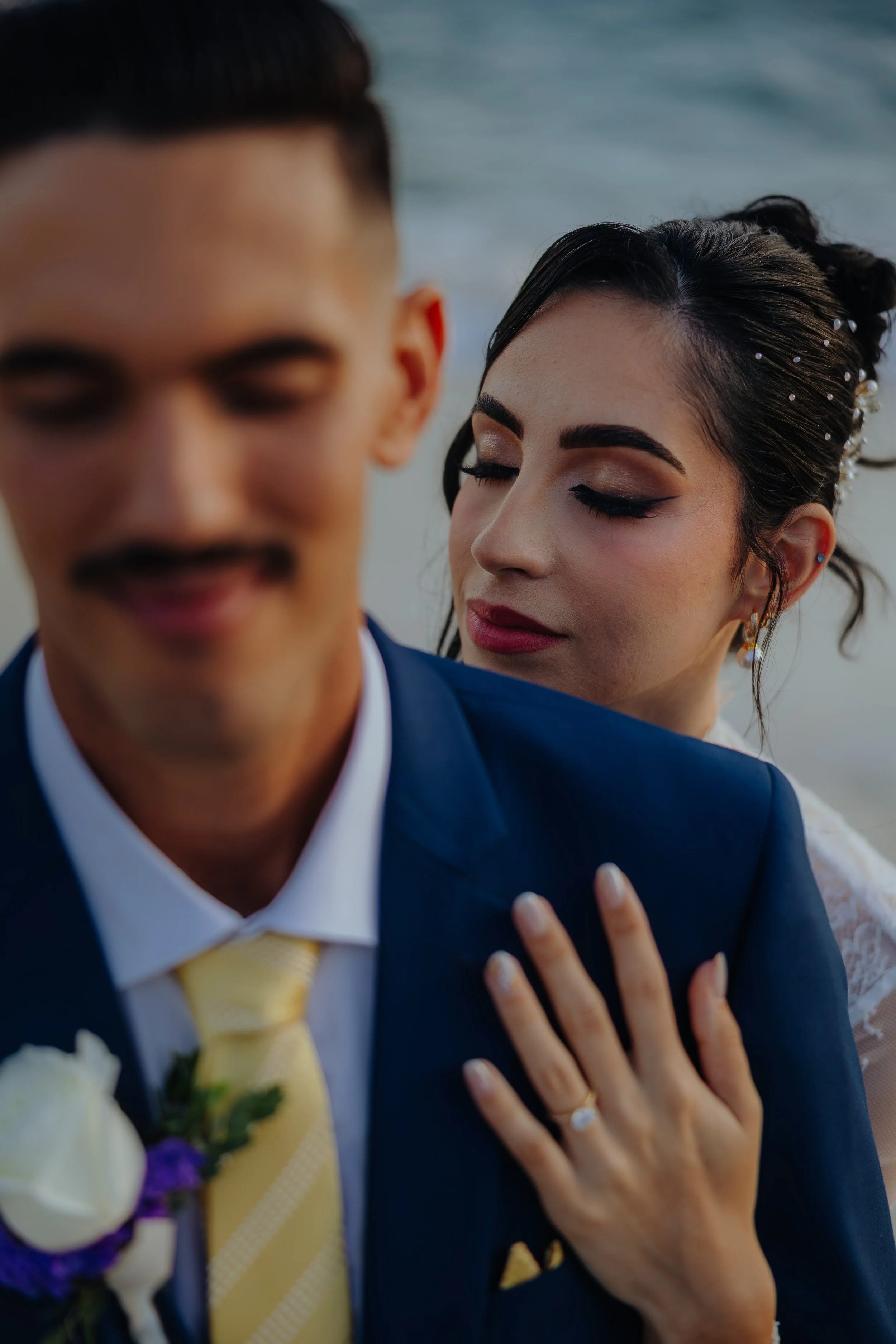 Close-up of a bride and groom at their wedding, with the bride resting her hand on the groom's chest, both dressed in formal wedding attire, with ocean in the background.