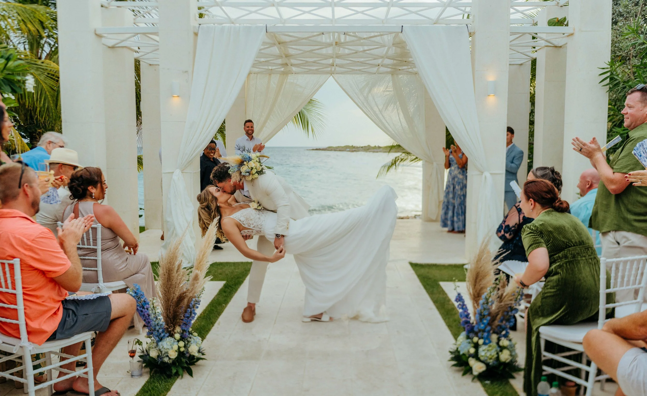 A wedding ceremony taking place outdoors at the beach, with a bride and groom sharing a kiss under a white canopy while guests watch and applaud.