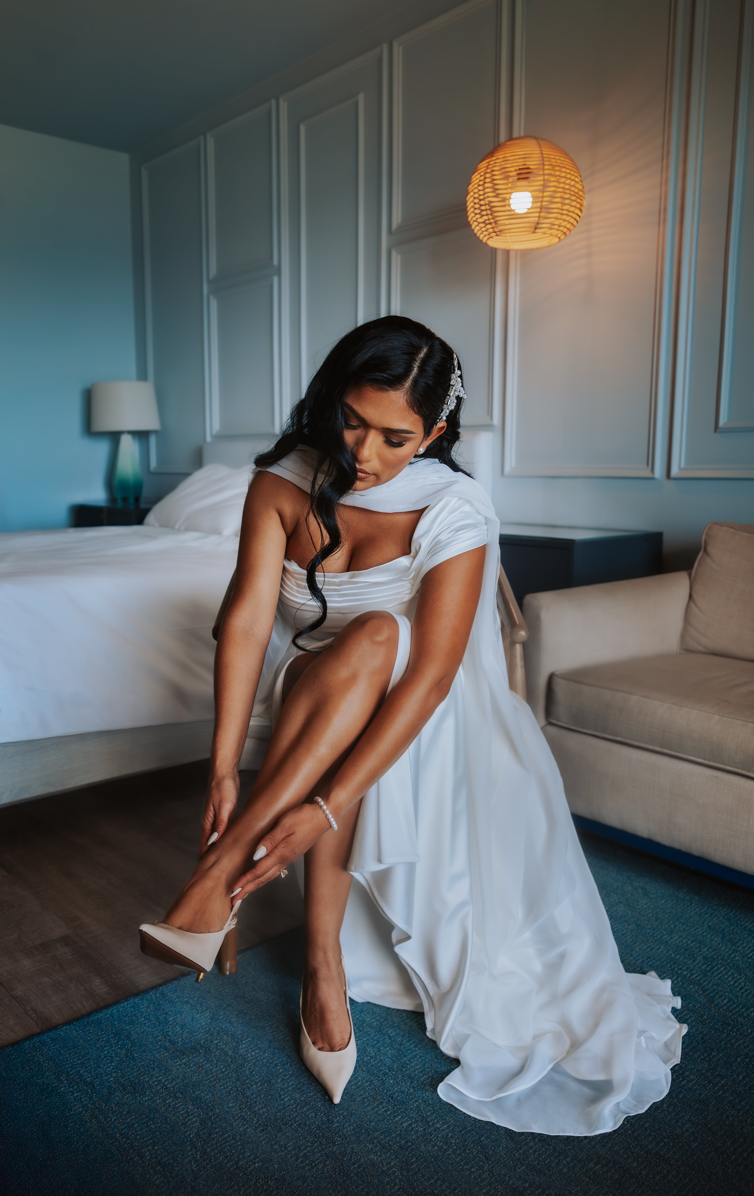 A bride with long dark hair, wearing a white wedding dress, getting ready and adjusting her high-heeled shoes in a hotel room with a bed, side table with lamp, and wall paneling