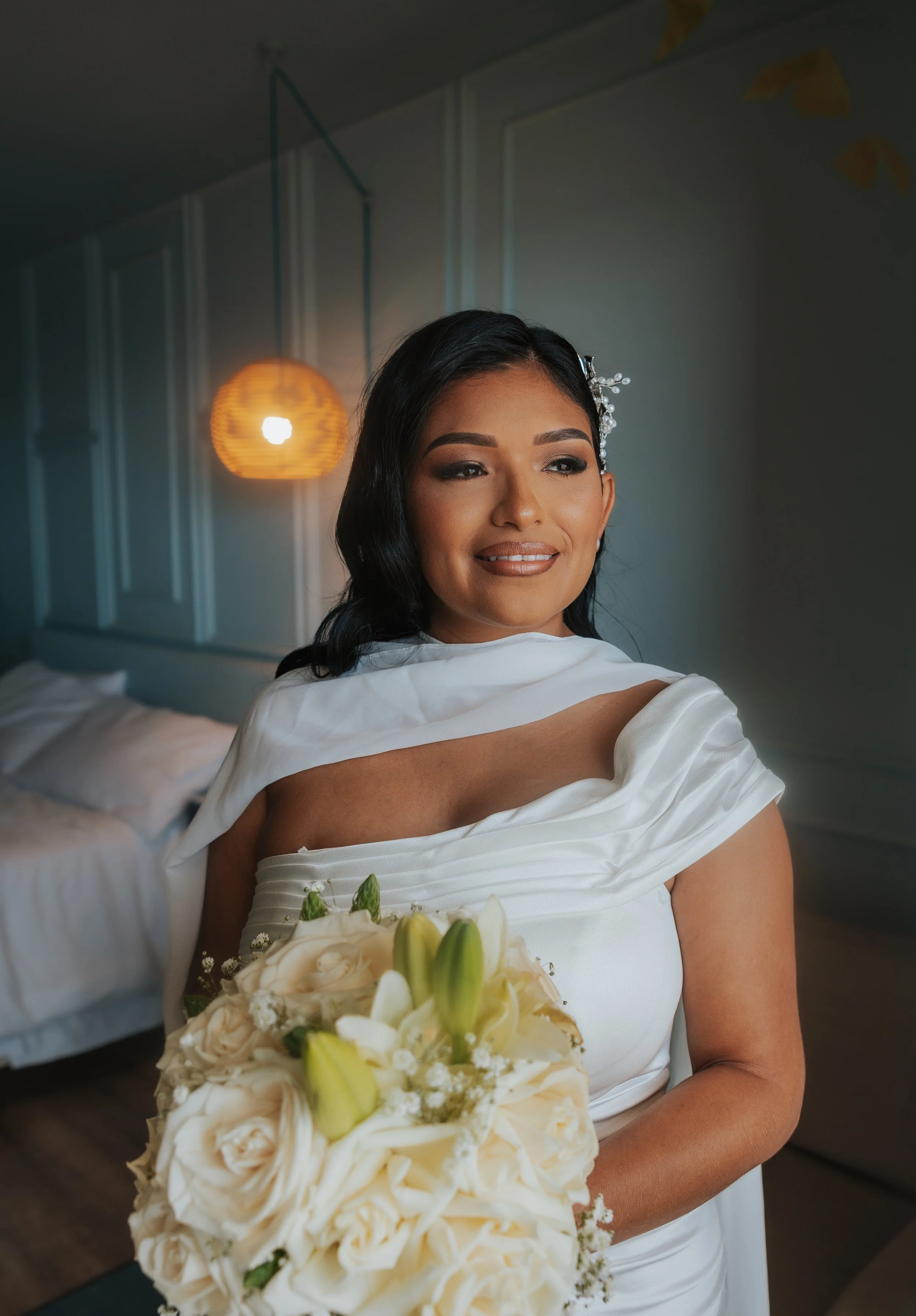 A woman in a white dress holding a bouquet of white flowers, standing in a room with a blue wall and a hanging orange lamp in the background.