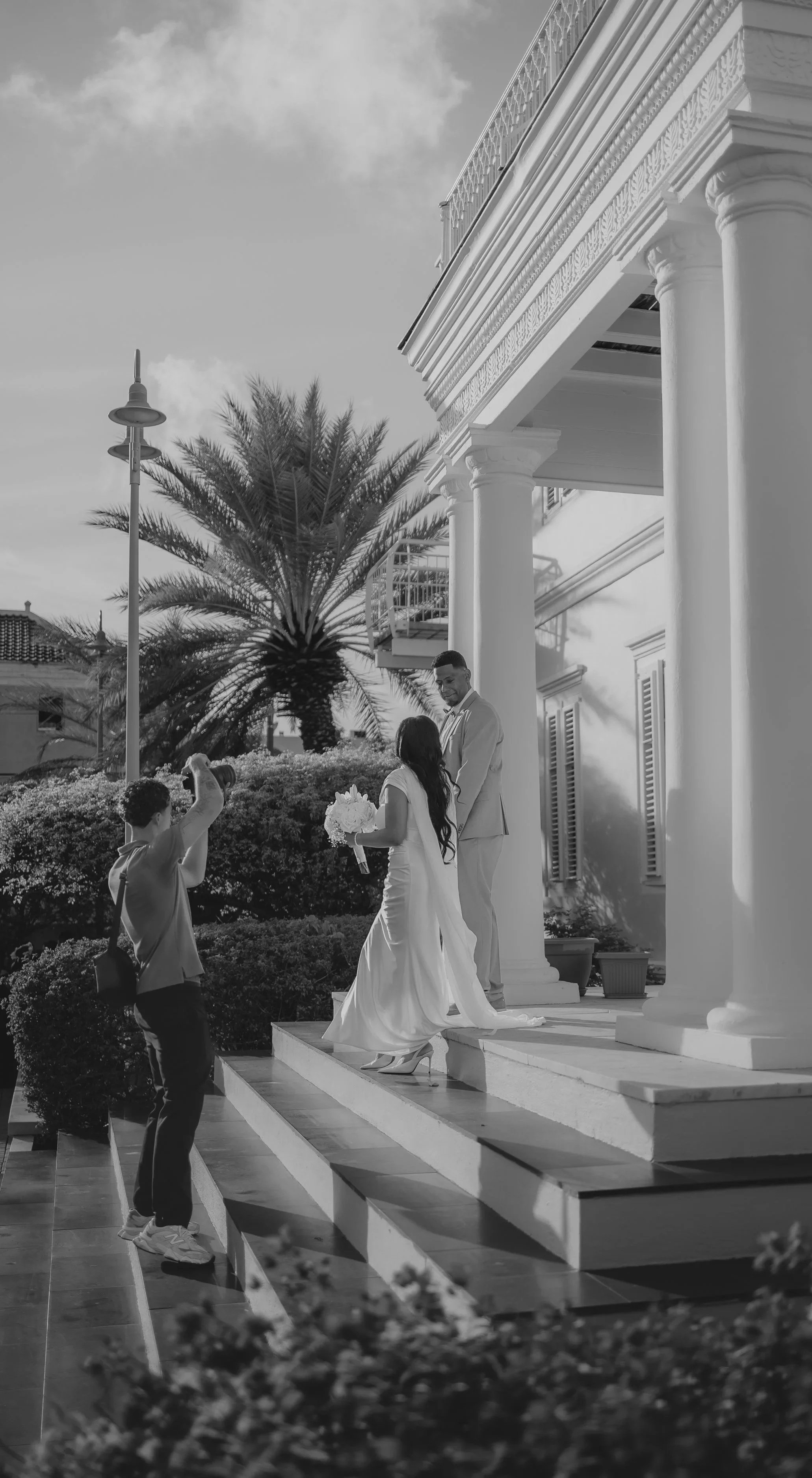 A wedding photo featuring a bride and groom on the steps of a white building with columns, with a photographer taking their picture, and a guest holding a bouquet of flowers. There are palm trees and a lamppost in the background.