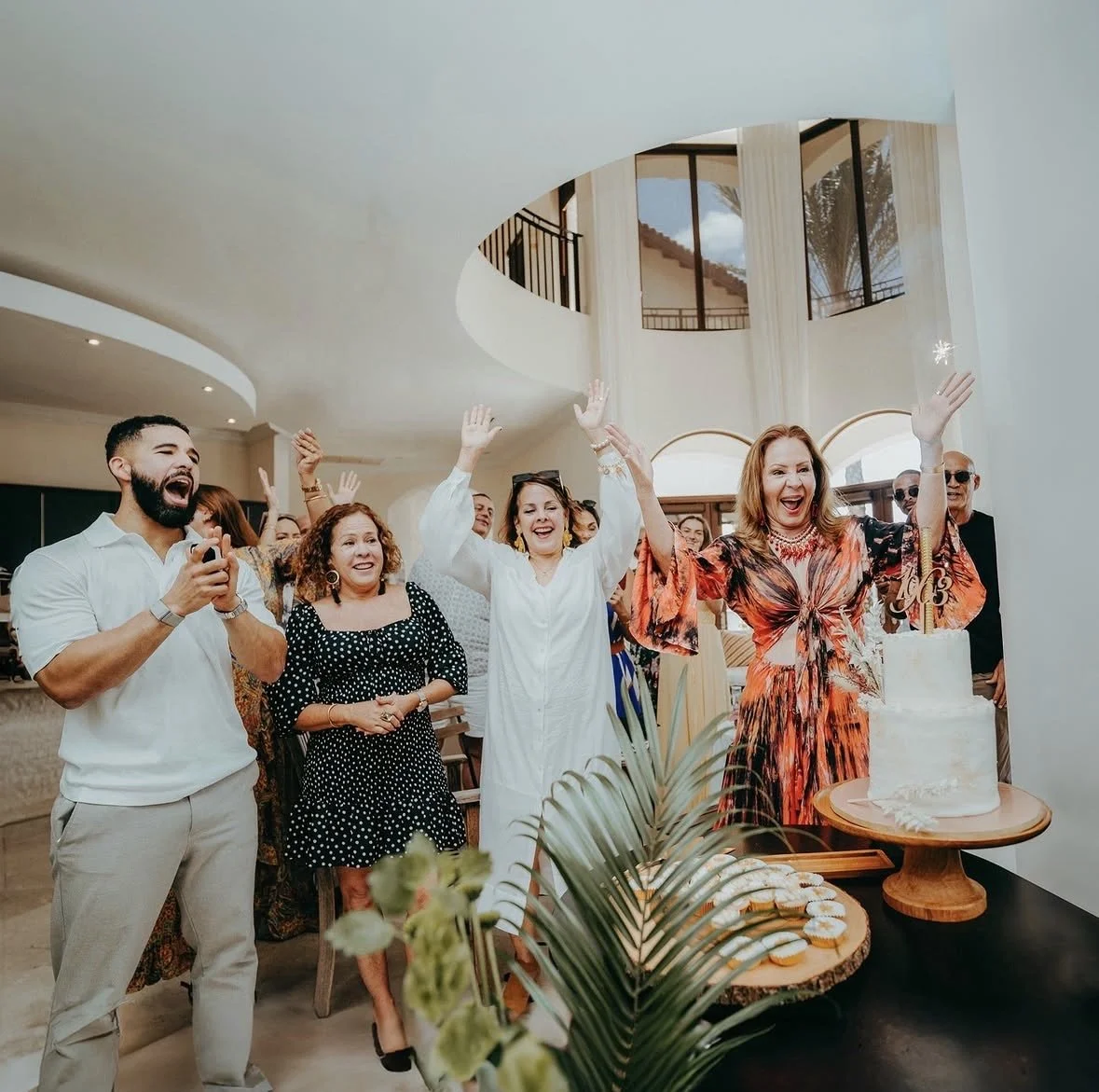 Group of people celebrating around a white layered cake with a gold monogram topper at a party or wedding reception indoors.