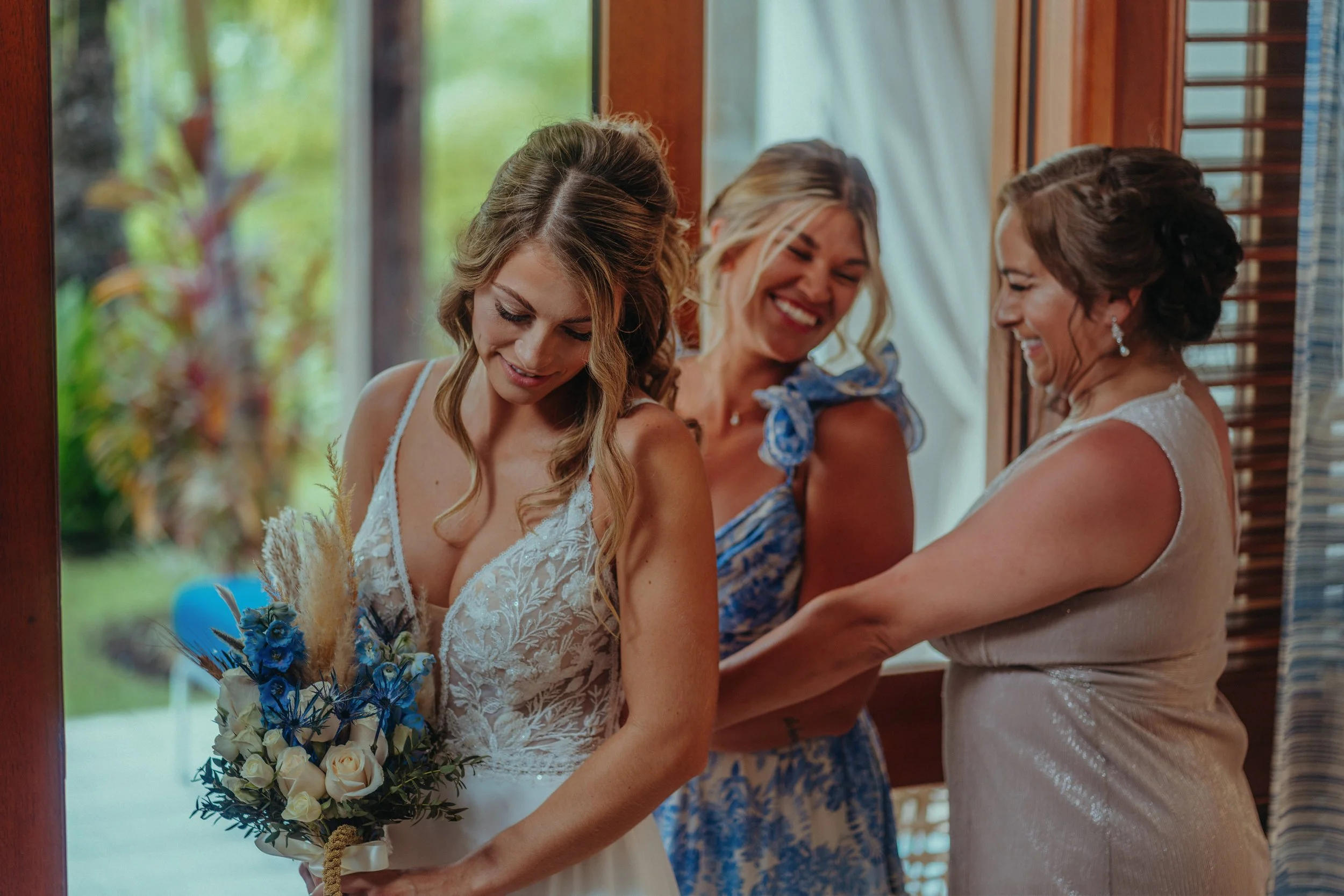 A bride in a white lace wedding dress holding a bouquet of blue, white, and beige flowers, standing with two women in a rustic setting with wooden walls and windows, smiling and sharing a moment.