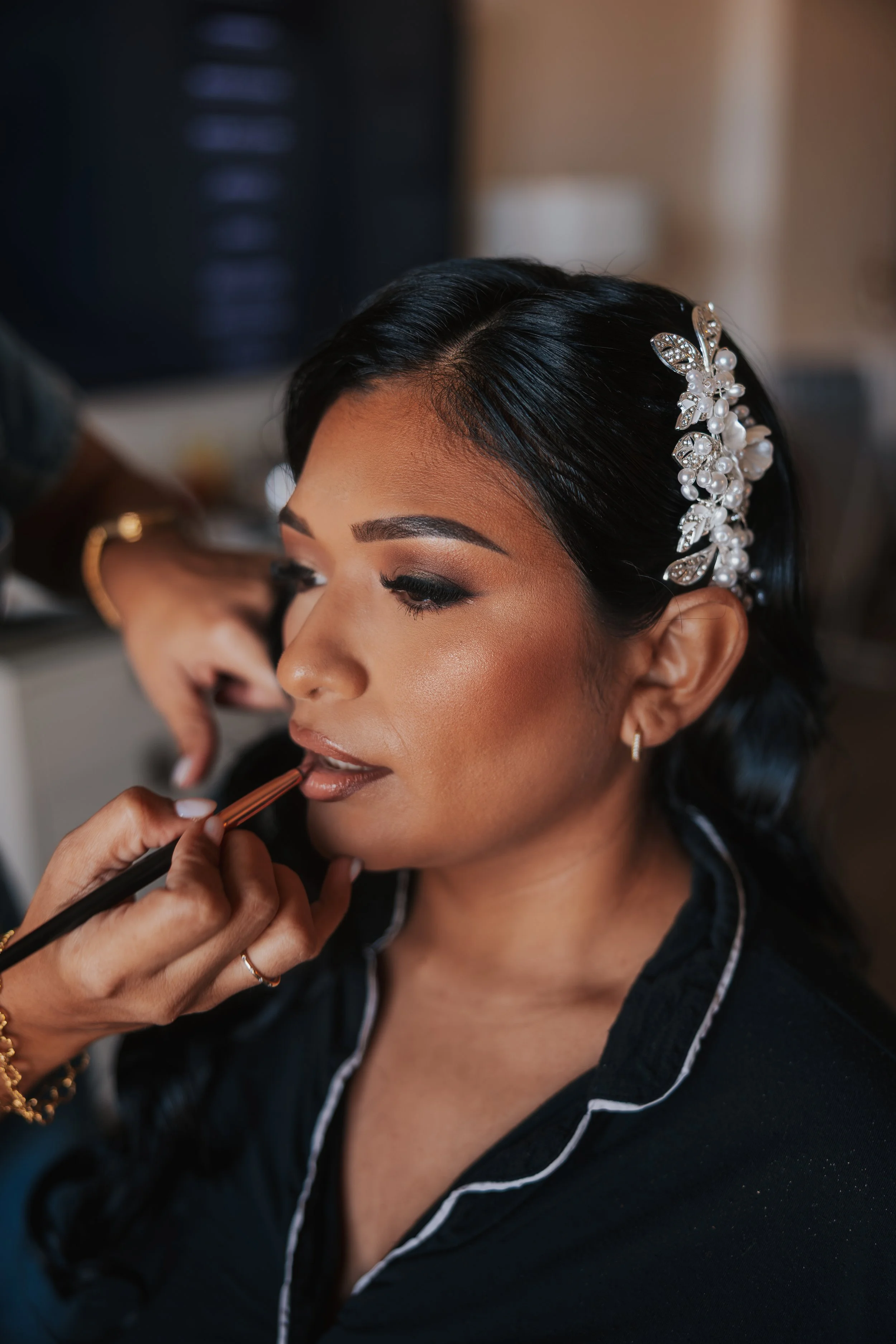 A woman getting her makeup done with a makeup artist applying lipstick. The woman has dark hair styled with an ornate pearl and jewel hairpiece, and she is wearing a dark top with white piping.