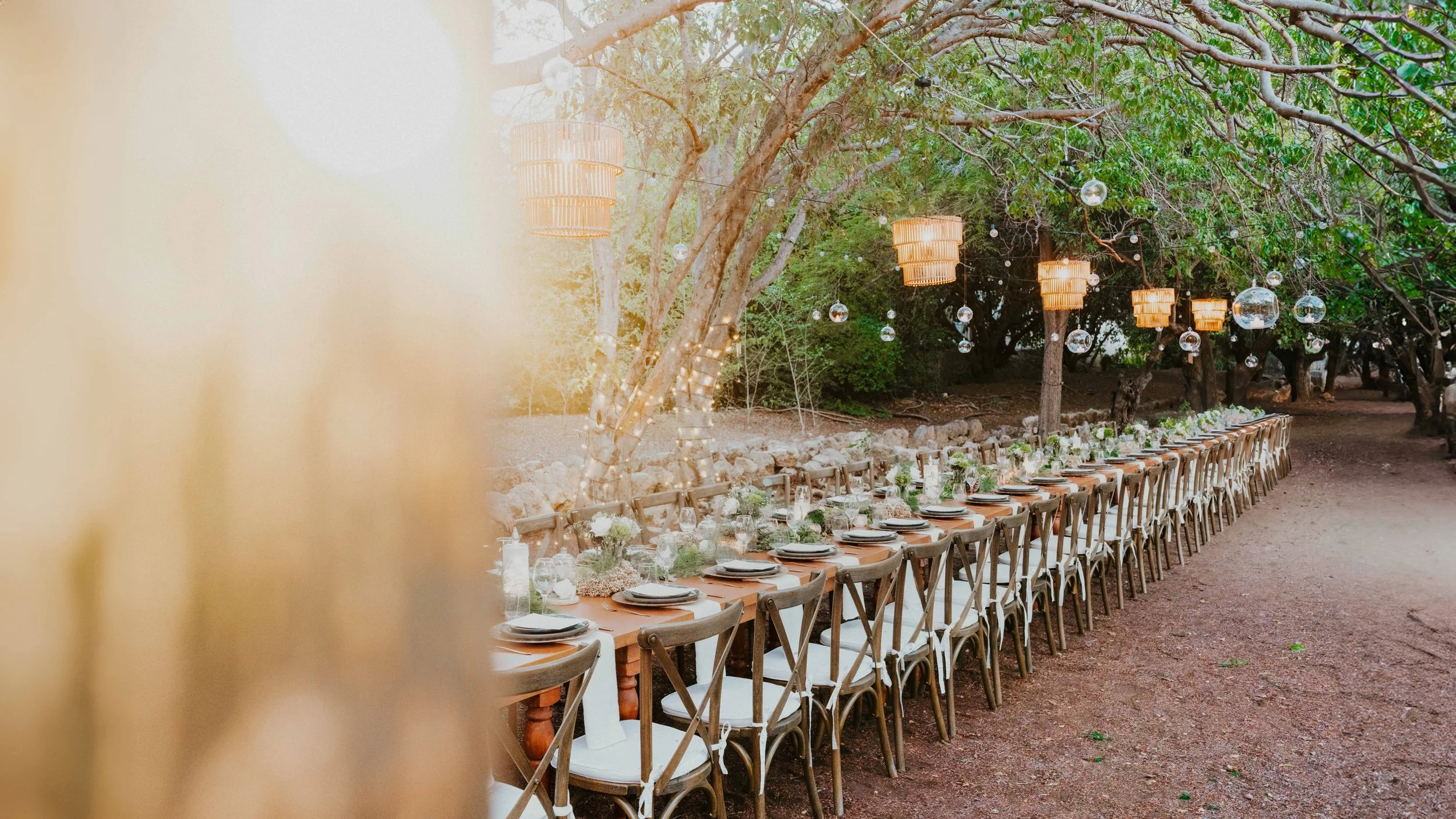 Long outdoor dining table set up for an event in a wooded area, decorated with floral arrangements, candles, and tableware, under hanging lantern-style lights.