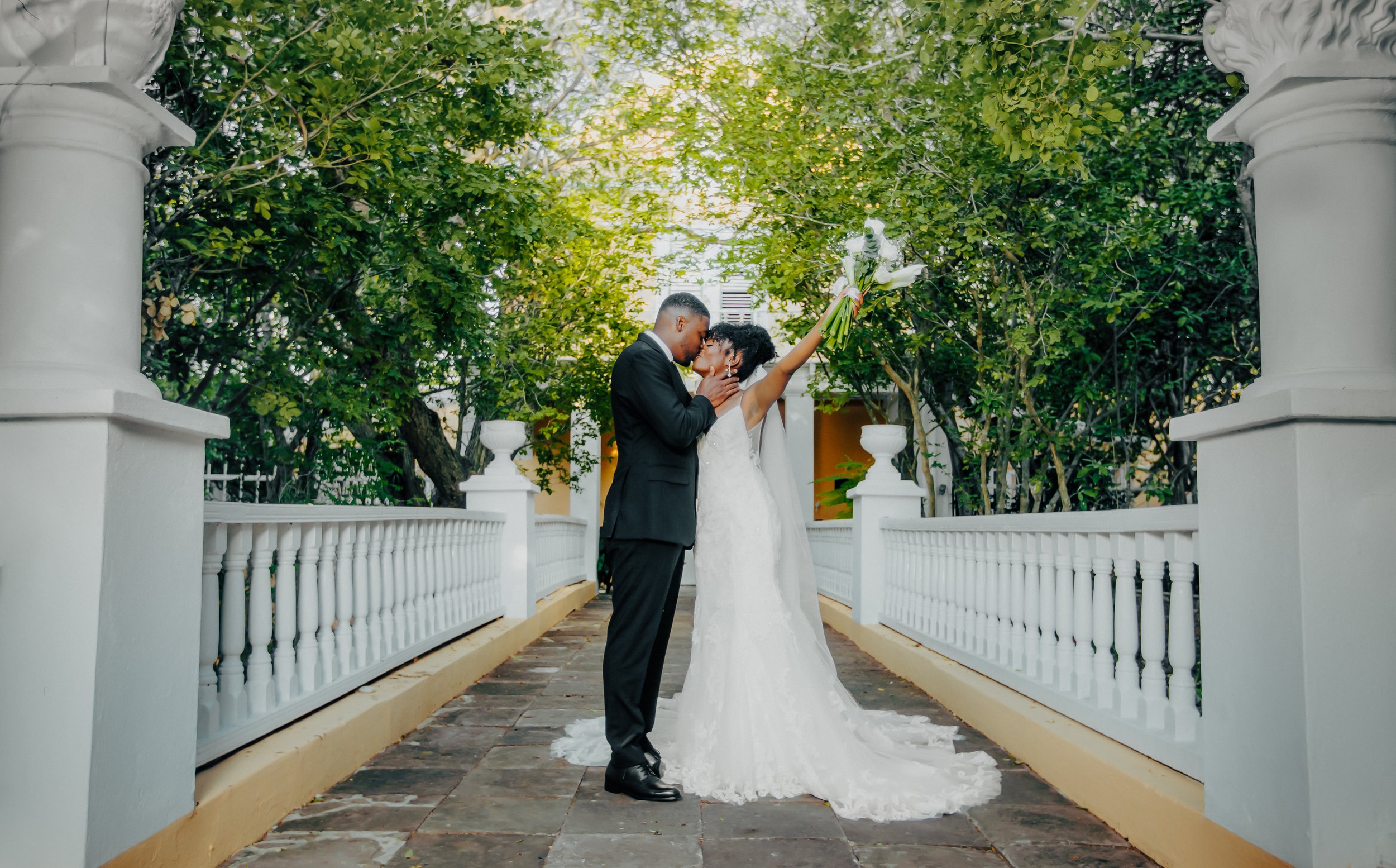 A bride and groom sharing a kiss on a small bridge surrounded by greenery at an outdoor wedding ceremony. The bride is holding a bouquet with her right hand raised in the air, while the groom is gently holding her face.