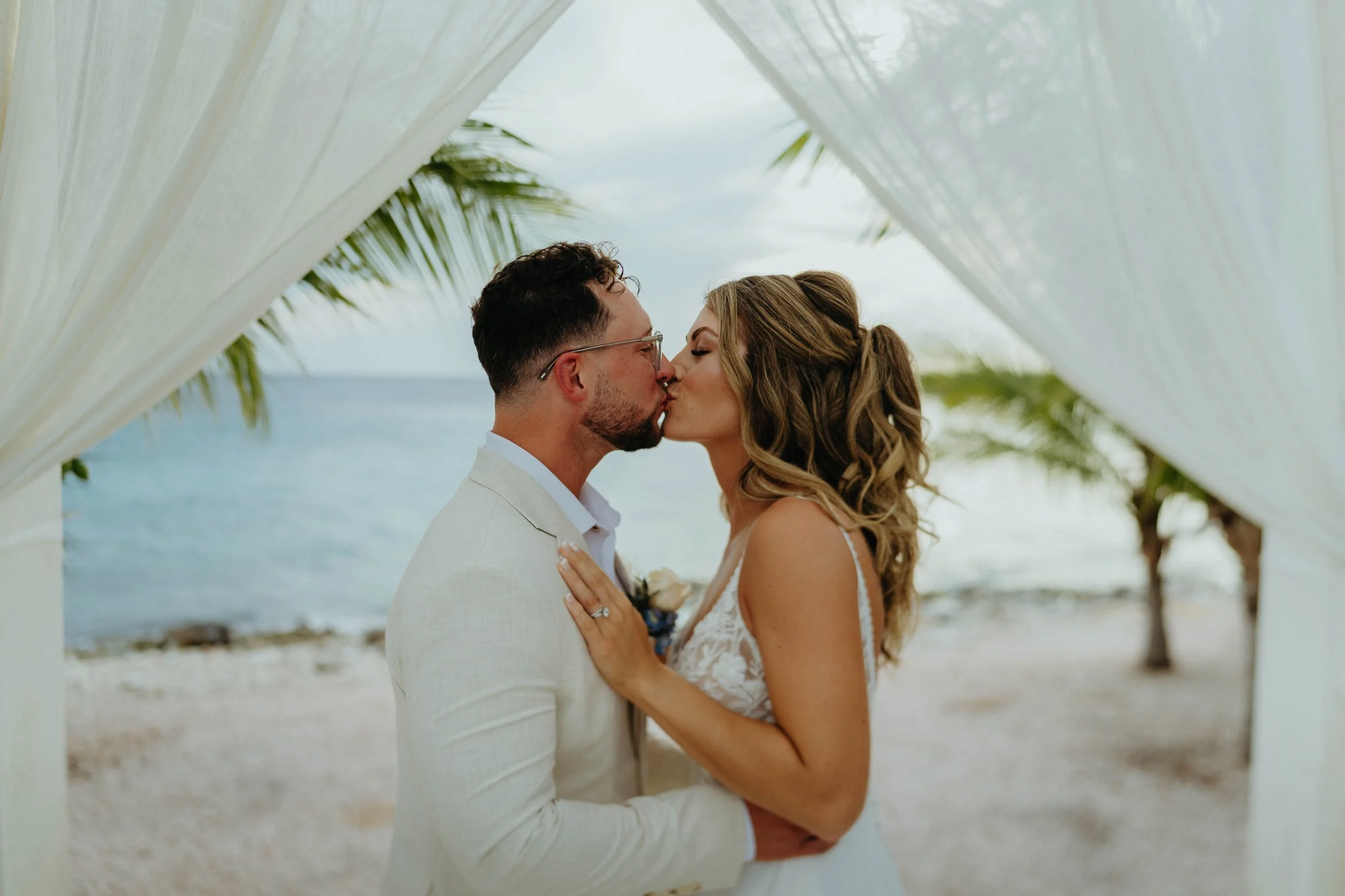 A couple sharing a kiss at the beach, framed by a white fabric canopy with palm trees and the ocean in the background.