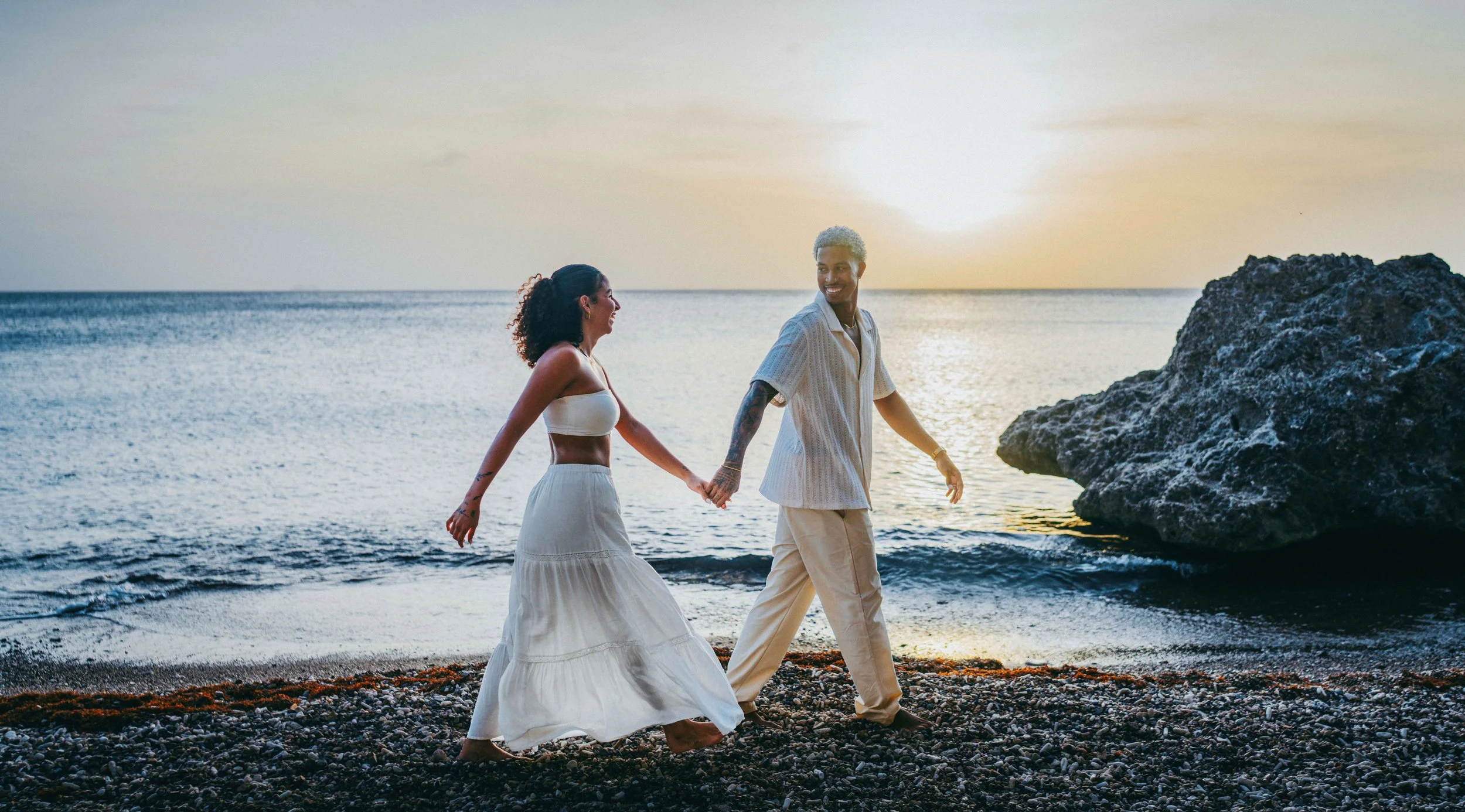 A happy couple holding hands and walking along the beach during sunset, with a large rock and the ocean in the background.