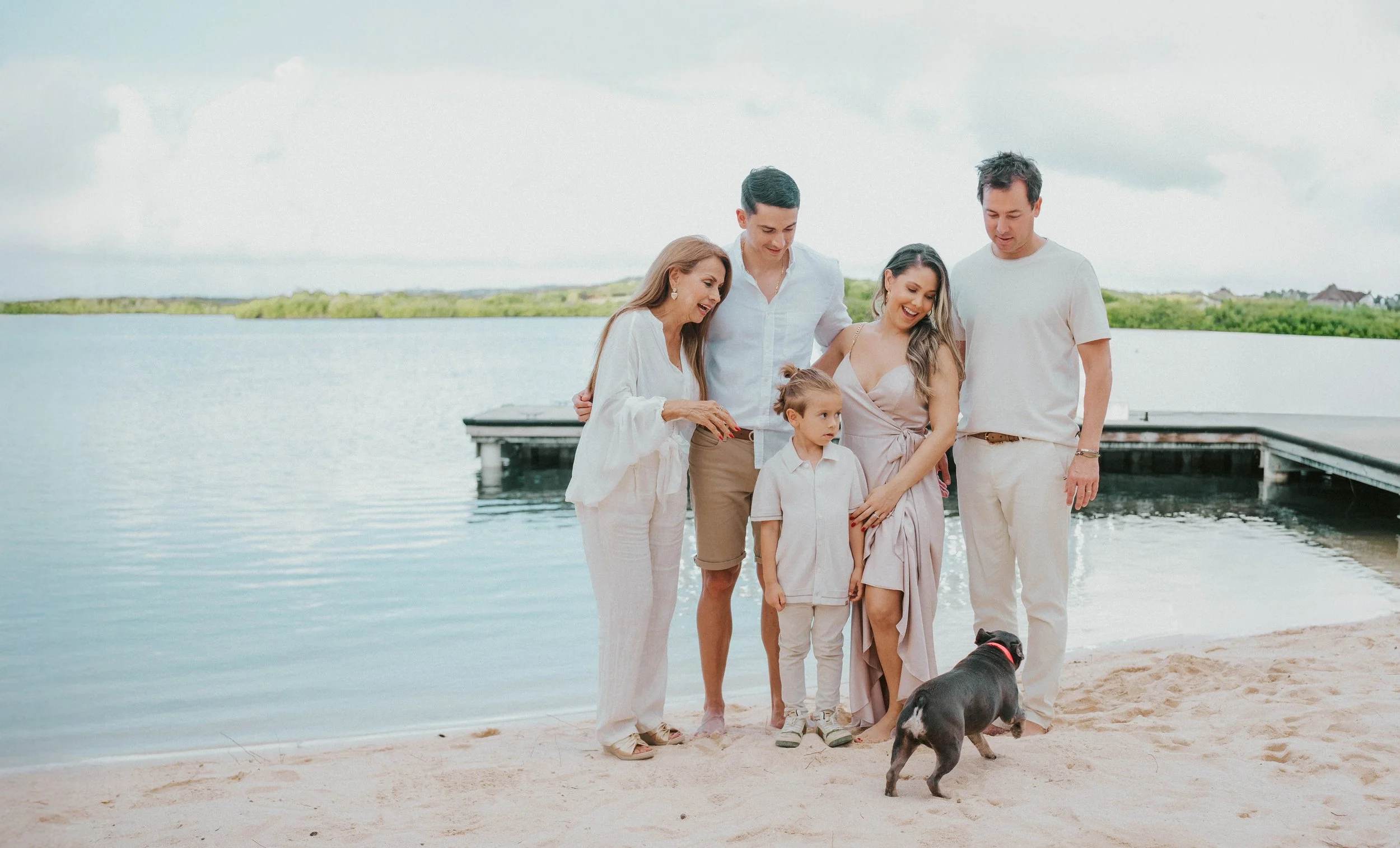A group of six people, including an older woman, three adults, and a young girl, standing on a beach near water, with a dog in the foreground, and a cloudy sky in the background.