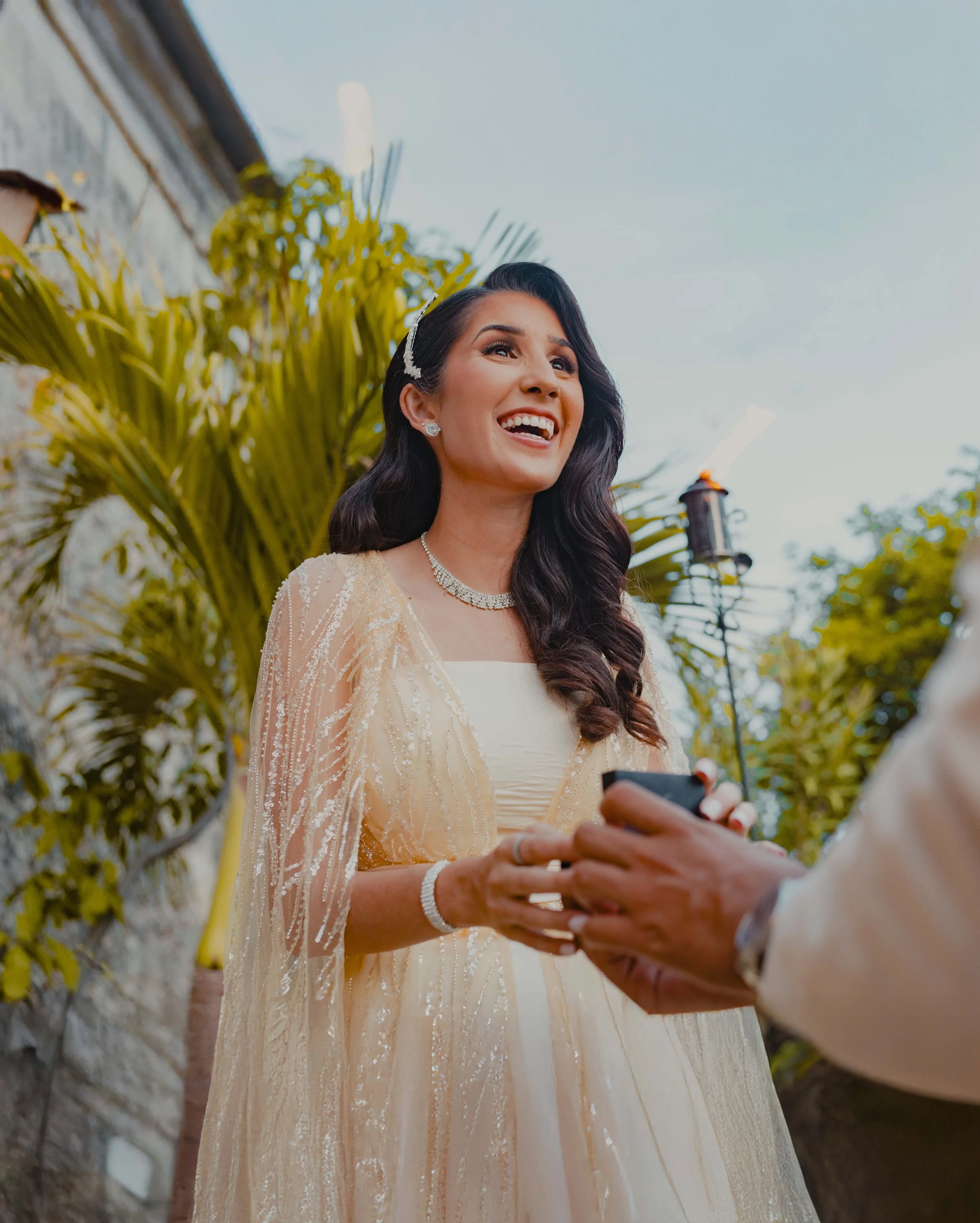 A smiling woman in a wedding dress and jewelry, holding hands with someone at an outdoor wedding ceremony.