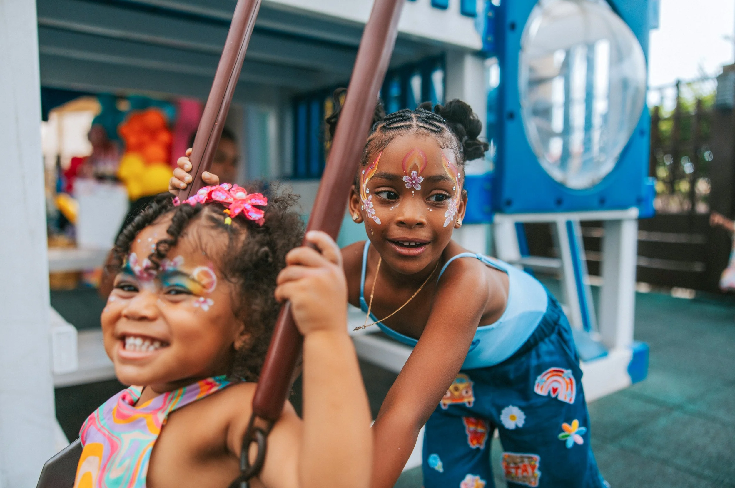 Two young girls are playing on a playground, with one girl doing a split while the other girl smiles and holds onto a playground bar. The girls have face paint and are smiling, enjoying outdoor activities.