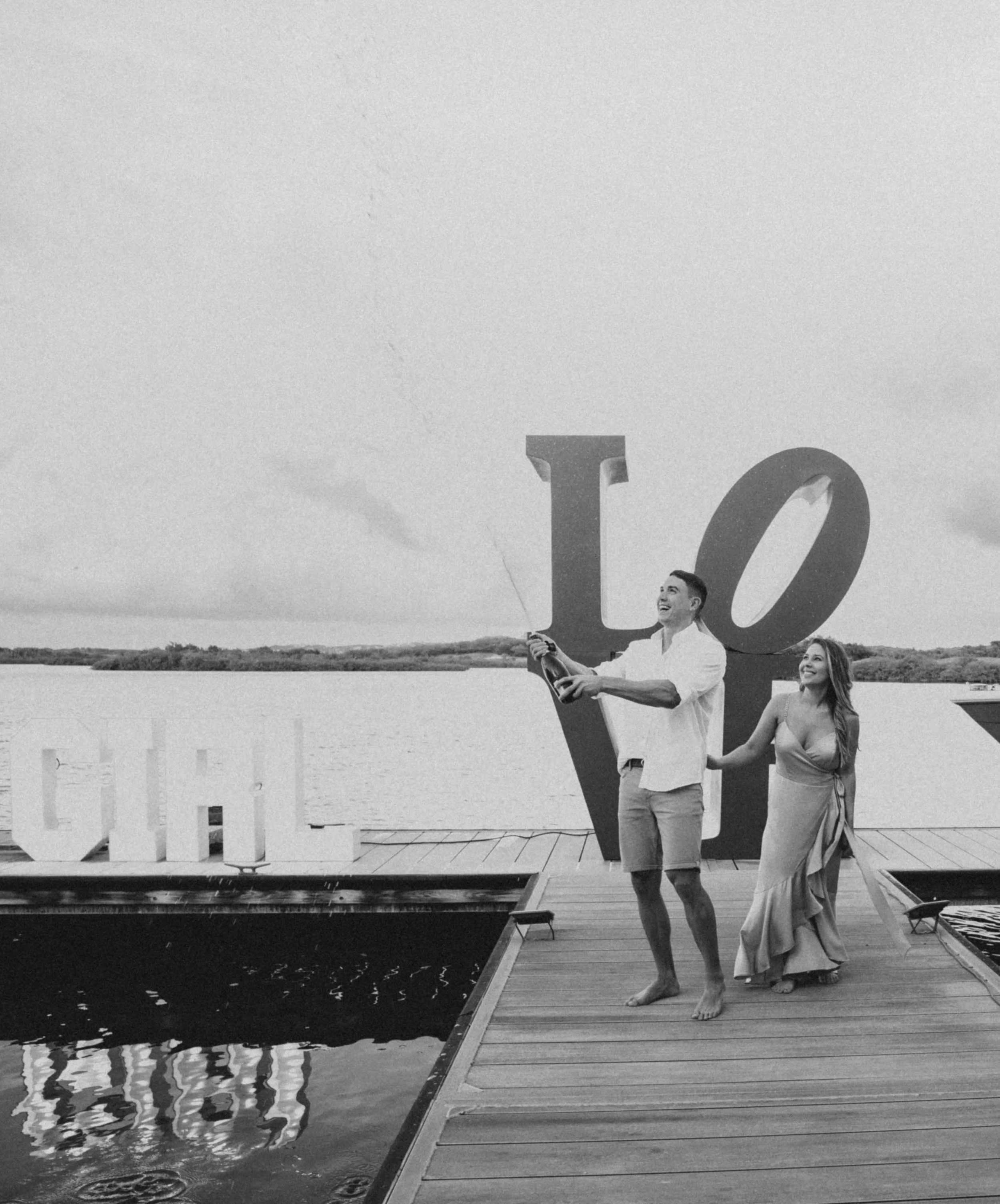 Two people, a man and a woman, walking along a dock next to a large decorative letter 'V' at a lakeside or river. The man is holding a bottle and is smiling as he looks ahead, while the woman is smiling and holding onto his arm. The sky is cloudy, an