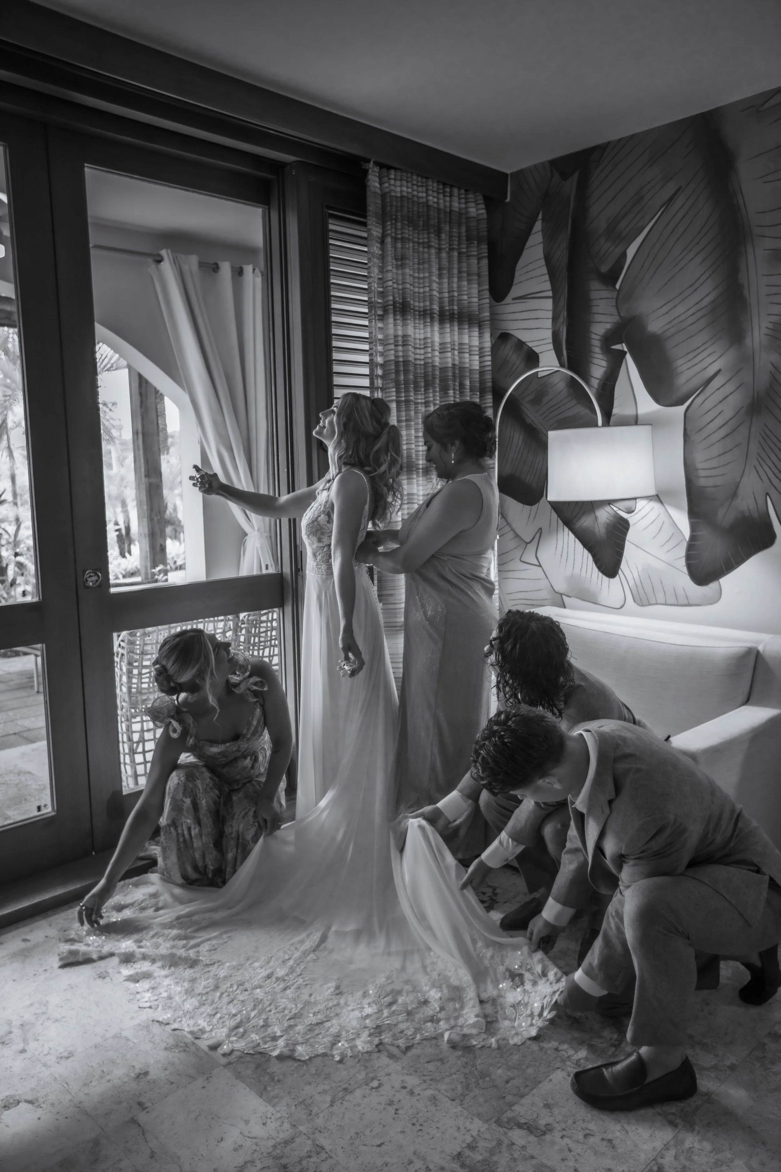 A bride in a wedding dress standing near a sliding glass door, surrounded by children and adults, as part of a wedding preparation or celebration inside a room with a large leafy mural on the wall and a lamp.