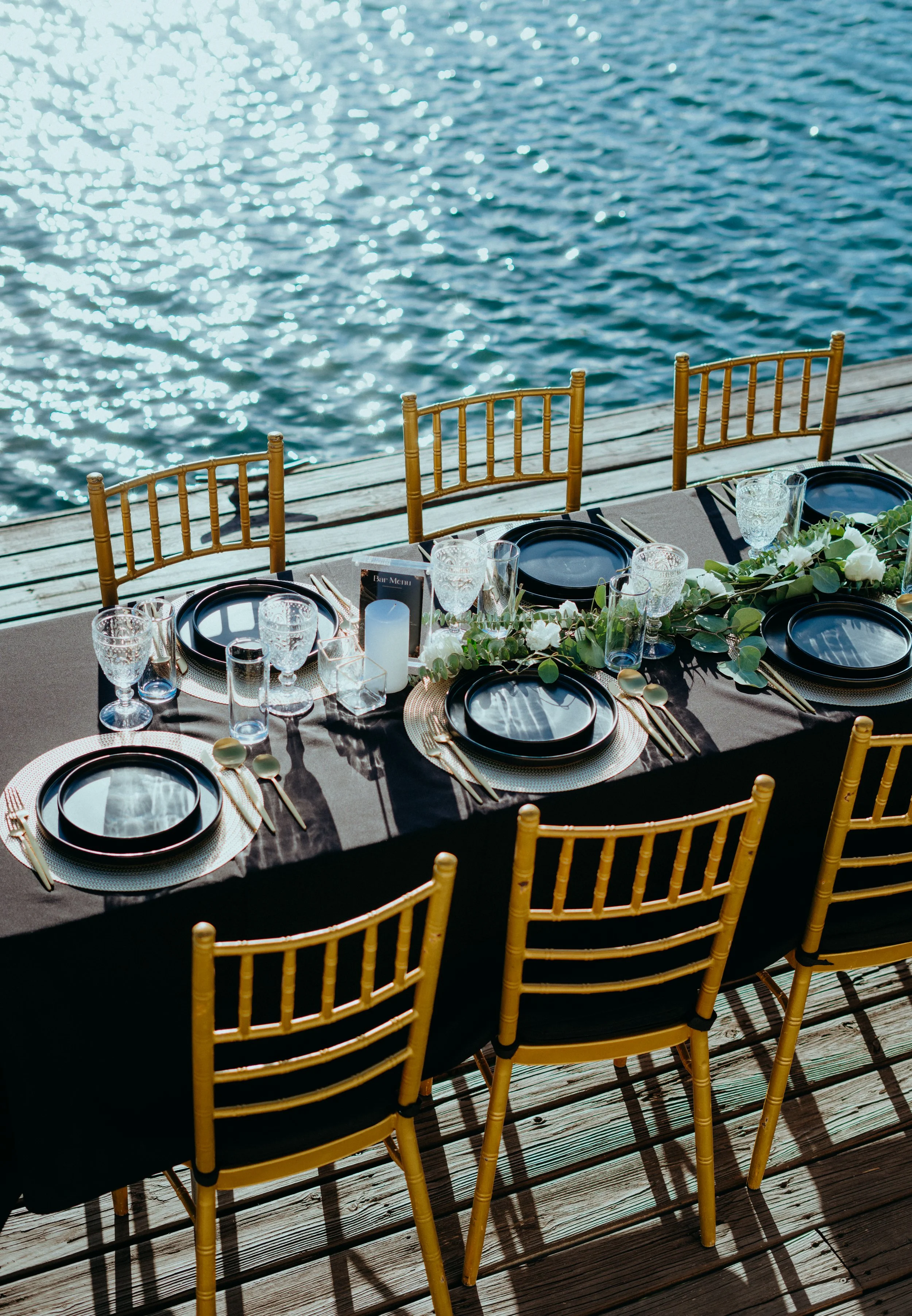 Outdoor dining table set on a wooden deck overlooking water with sunlight reflections, decorated with black plates, glassware, a floral centerpiece, and gold chairs.