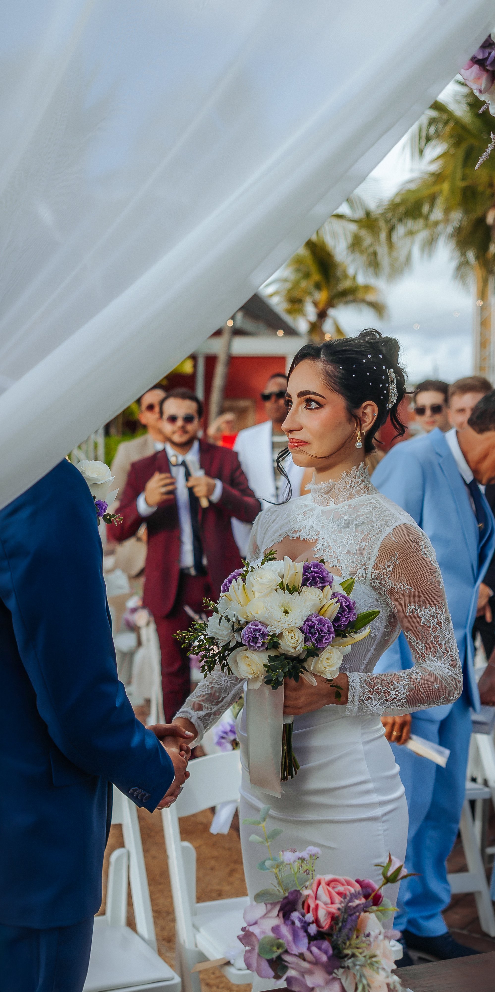 A bride holding a bouquet of white and purple flowers during a wedding ceremony, facing a groom in a blue suit, with guests in formal attire in the background under a partly cloudy sky and palm trees.