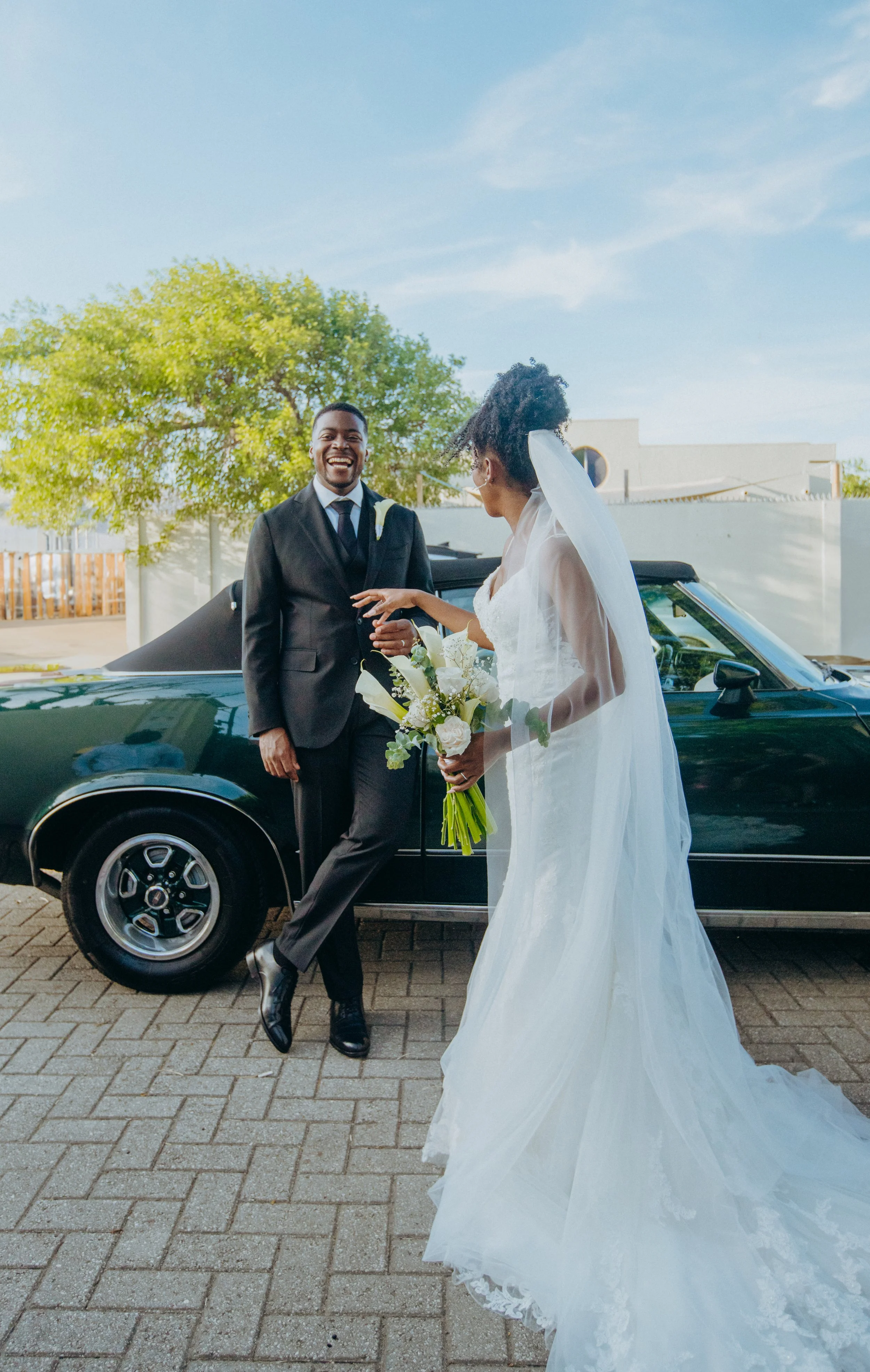 A bride in a white wedding gown and veil holding a bouquet of white roses and greenery smiling as a groom in a black suit and tie stands next to her, both smiling, with a vintage black car and a tree in the background outdoors on a sunny day.