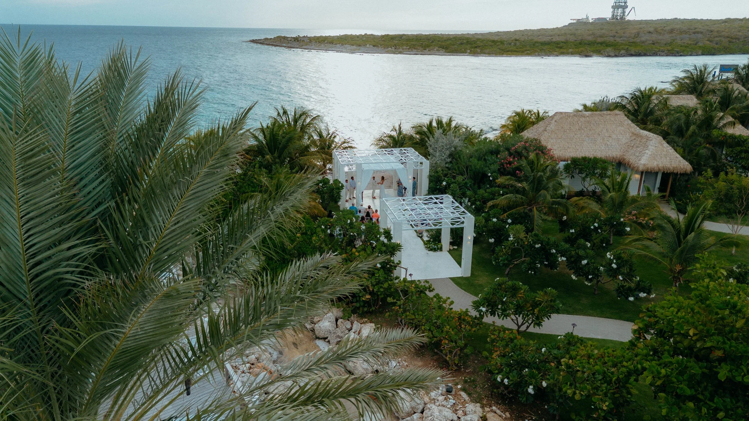 Beachside wedding ceremony setup with white structures, surrounded by lush green foliage and overlooking the ocean.