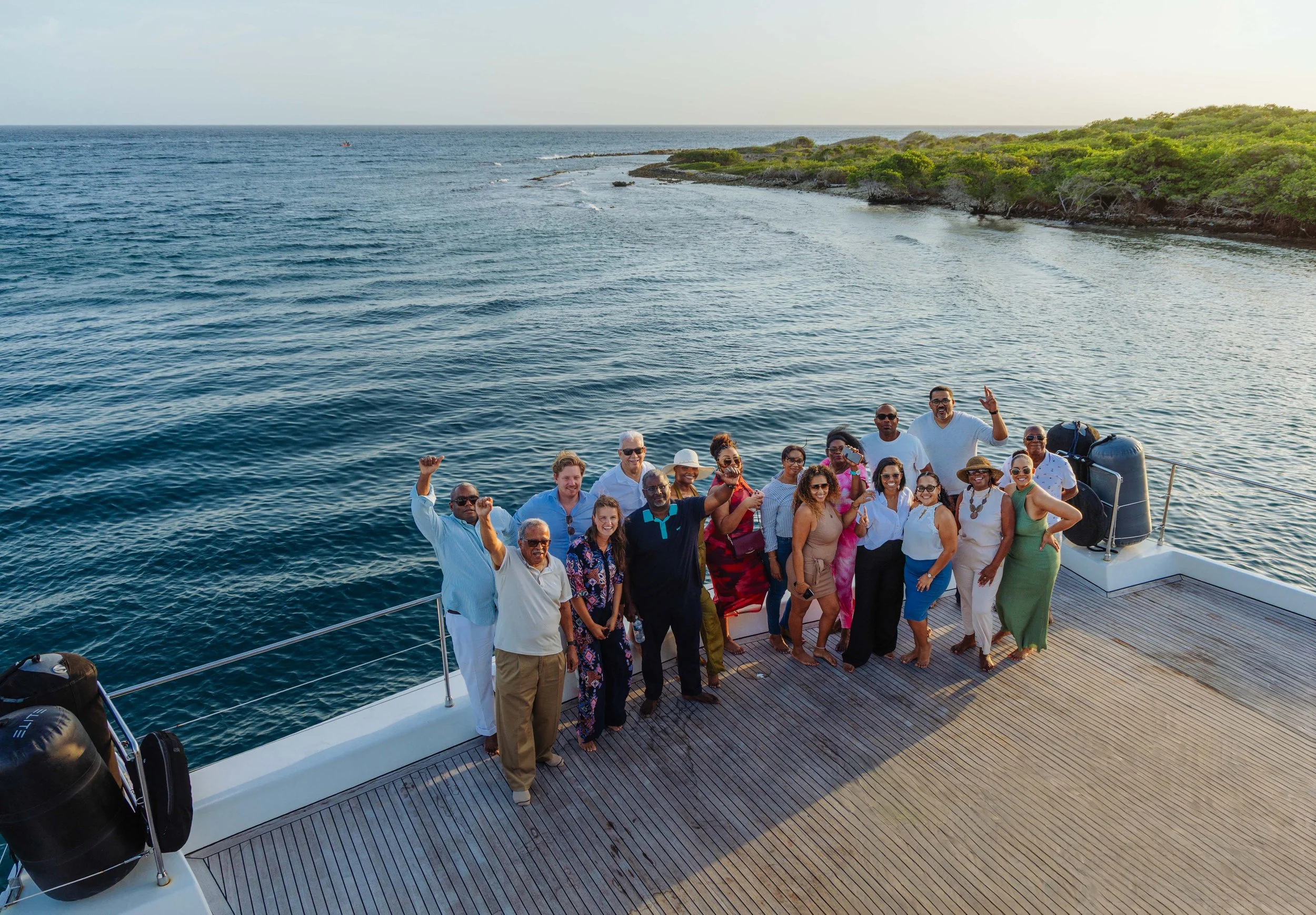 A group of people on a boat deck near a lush, green coastline under a clear sky, enjoying a sunny day.