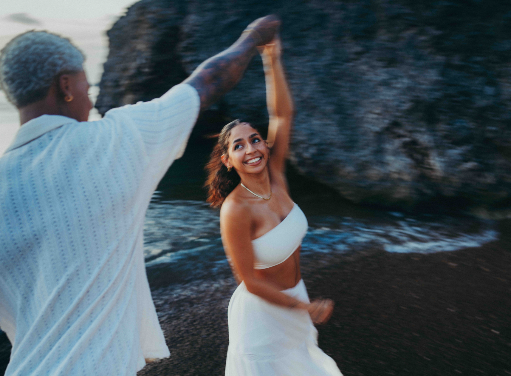A young woman with medium skin tone, wearing a white strapless top and skirt, smiling and dancing on a shoreline near rocks, with a man in a white shirt with tattoos on his arm holding her hand and leading her in dance.