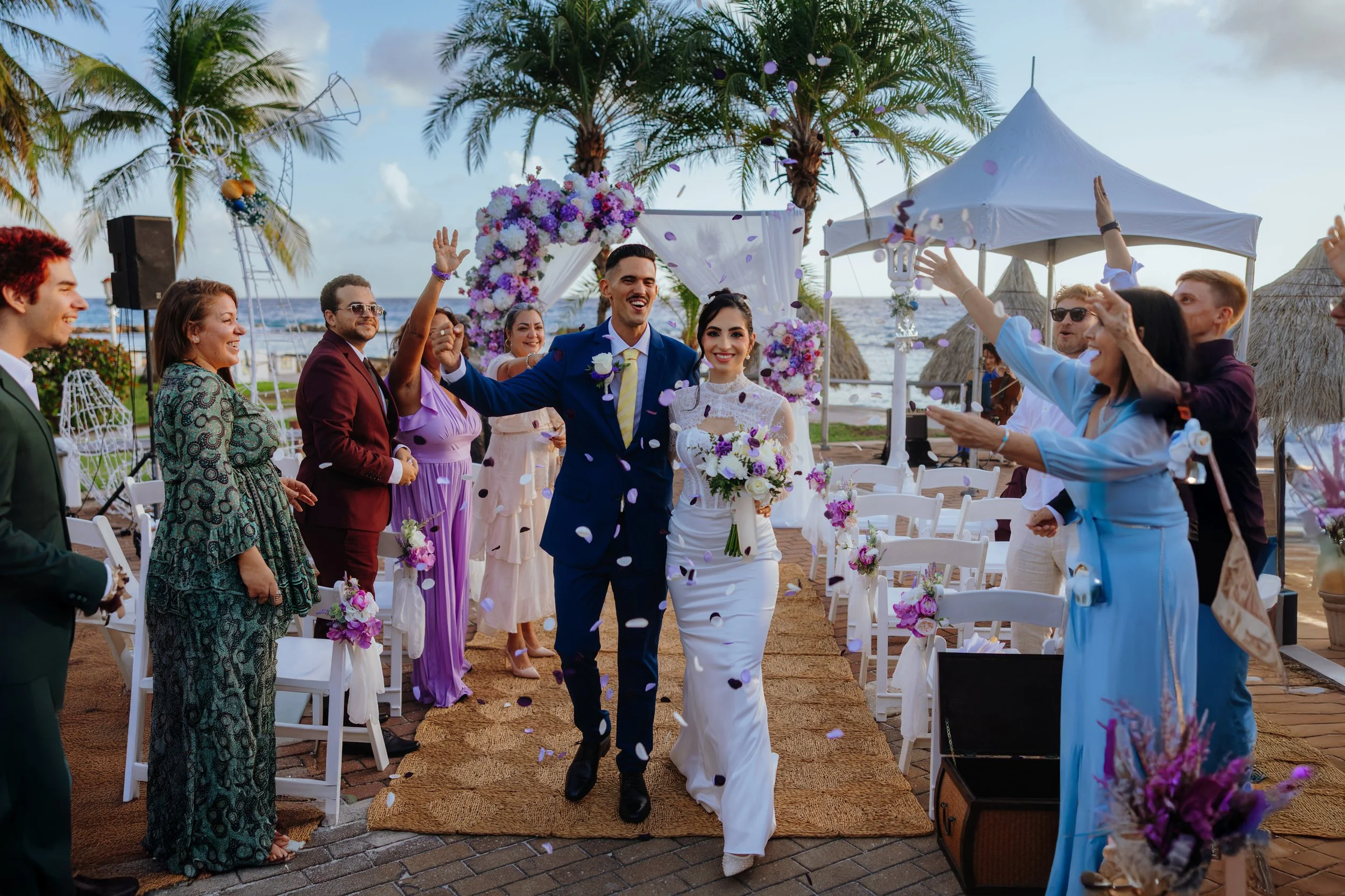 A bride and groom walking down the aisle at an outdoor beach wedding, surrounded by friends and family celebrating with confetti and smiles.