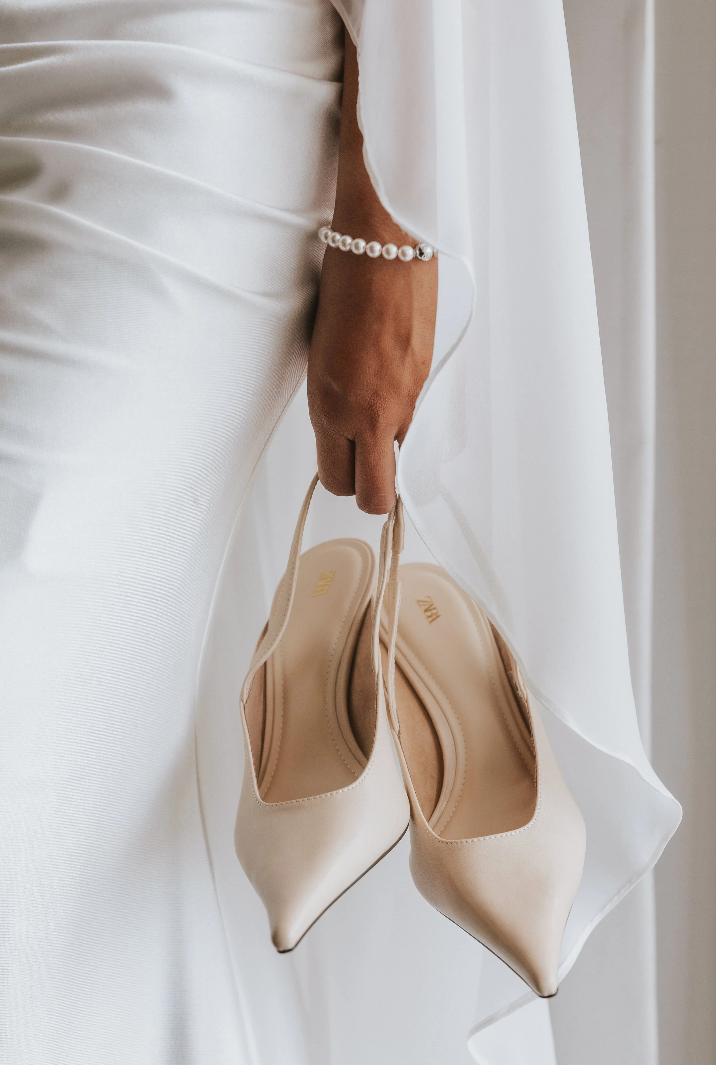 A pair of beige high-heeled shoes hanging from a person's hand, with a white dress or fabric in the background, and the person wearing a pearl bracelet.