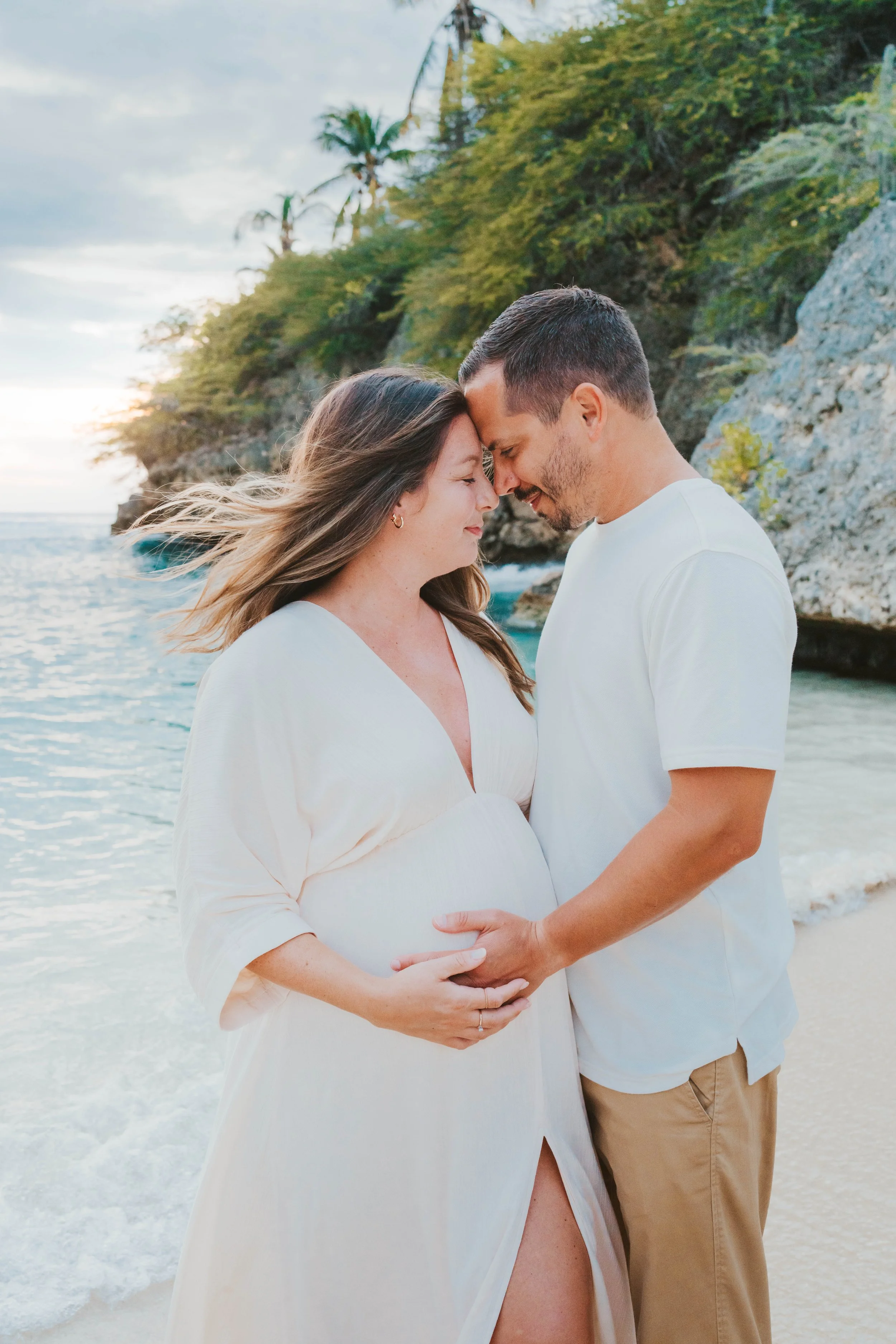 A pregnant woman and a man standing close together on a beach with rocky cliffs and palm trees in the background. They are touching foreheads and smiling, with the woman's hand on her belly.