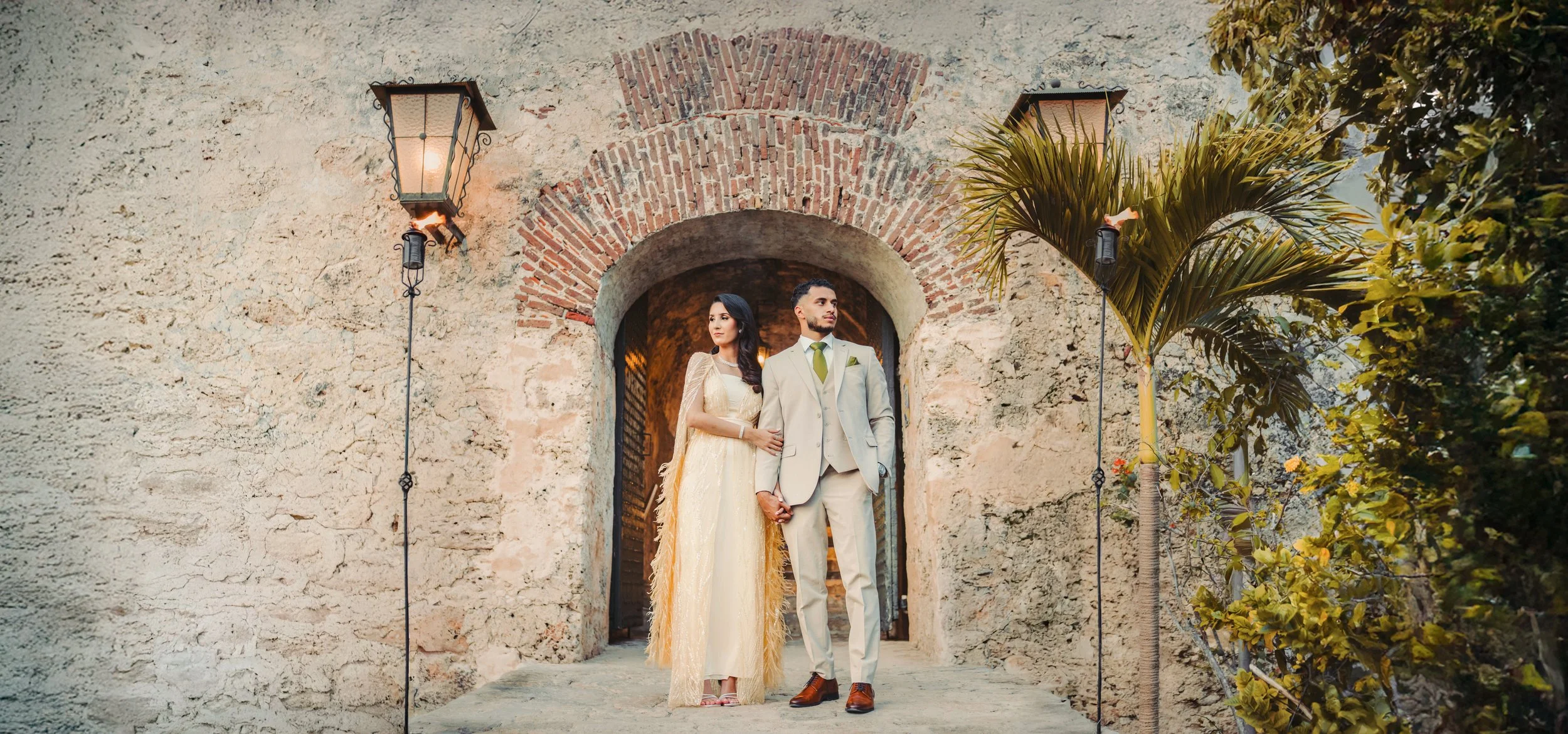 A bride and groom standing hand in hand in front of an arched doorway made of stone and brick, with vintage street lamps on either side and greenery around.