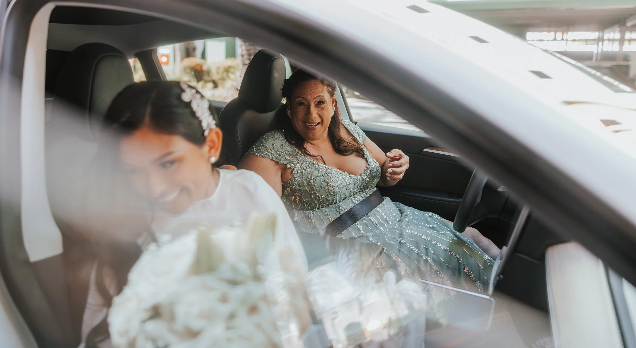 Women wearing formal dresses sitting inside a vehicle, with the woman in the background smiling and talking, seen through the windshield.