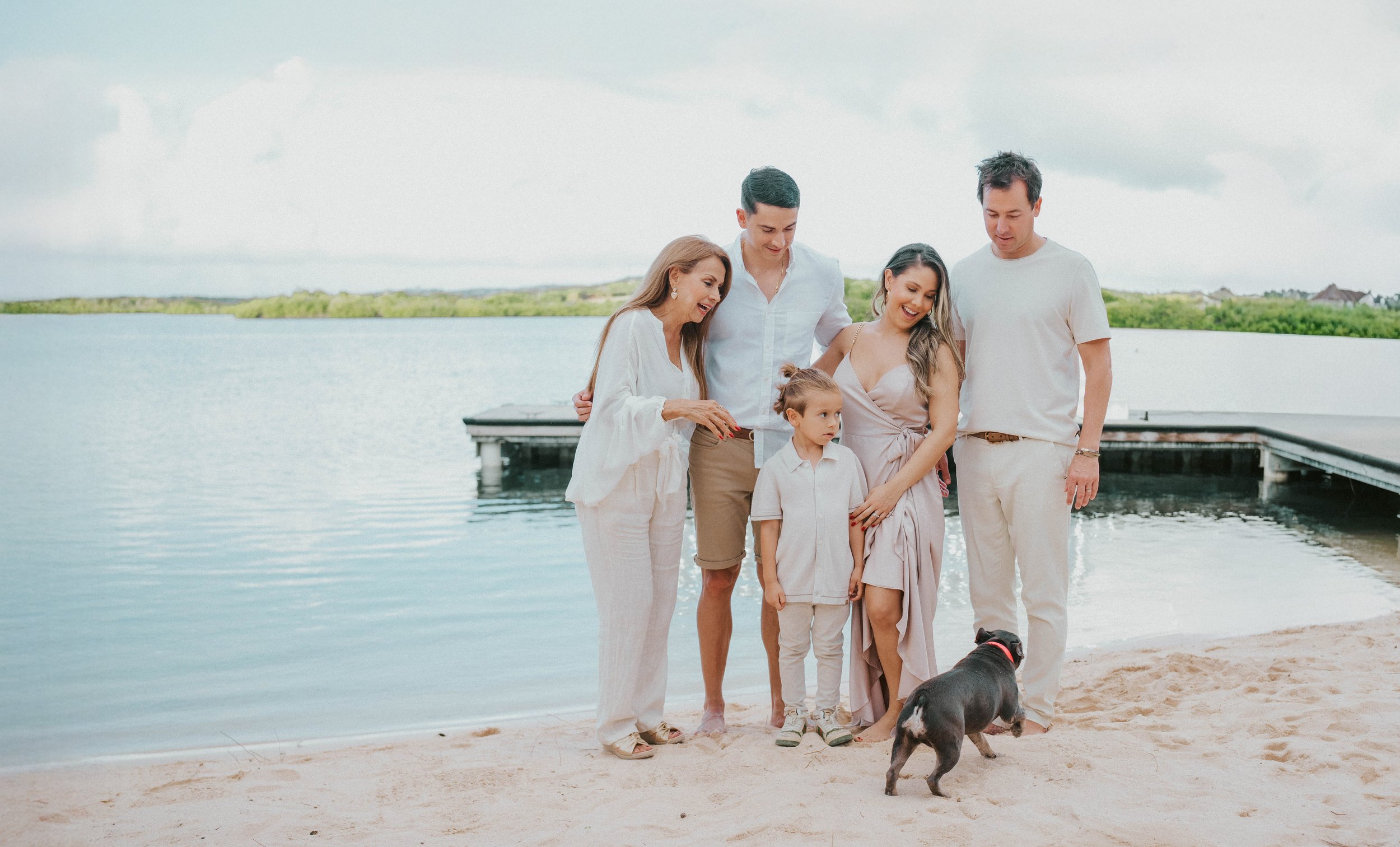 A family of six, including two children, stands on a sandy beach near a lake with a dock, some houses, and greenery in the background, smiling and looking at a dog.