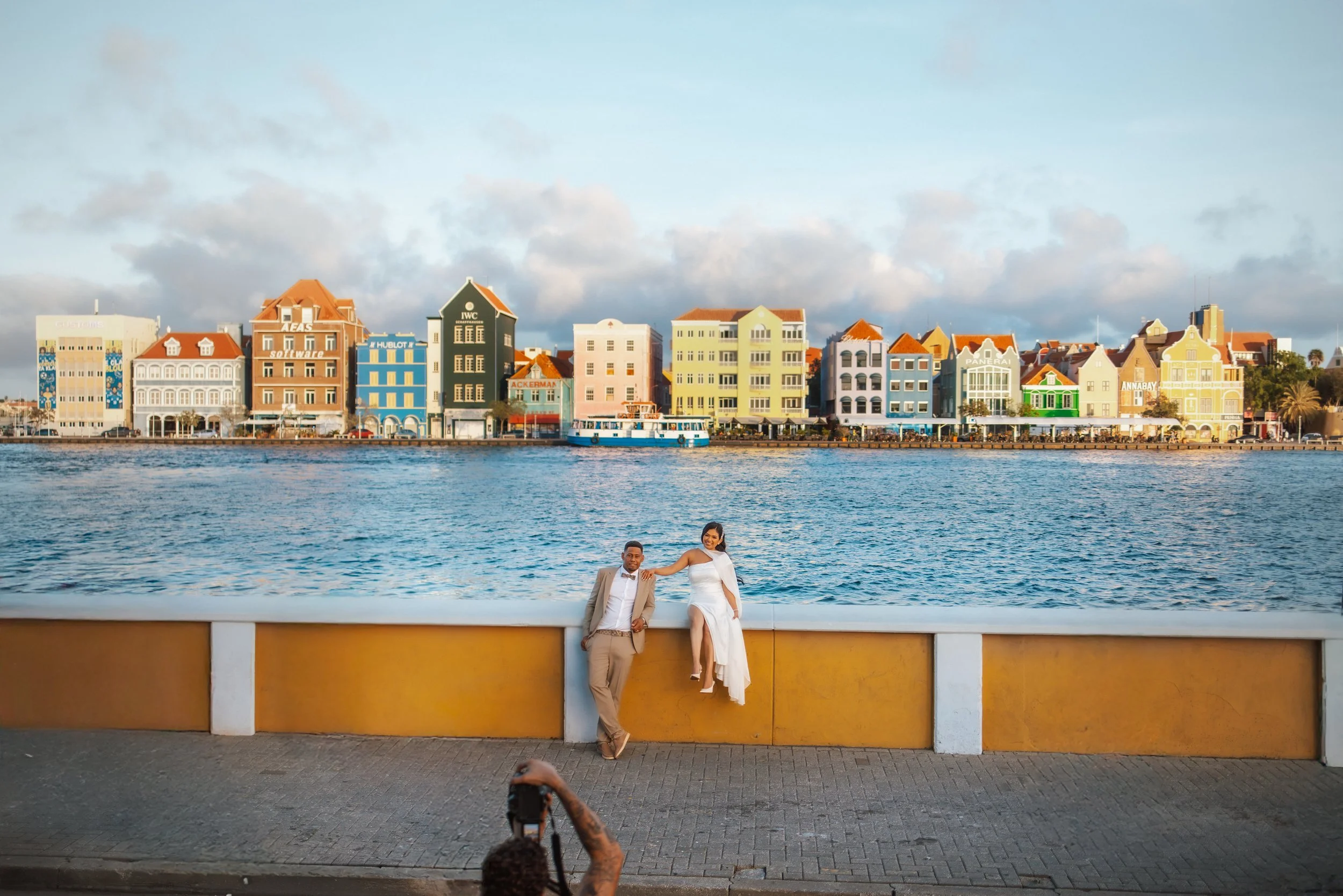 A couple dressed in formal attire standing on a waterfront walkway with colorful buildings across the water and a cloudy sky in the background.