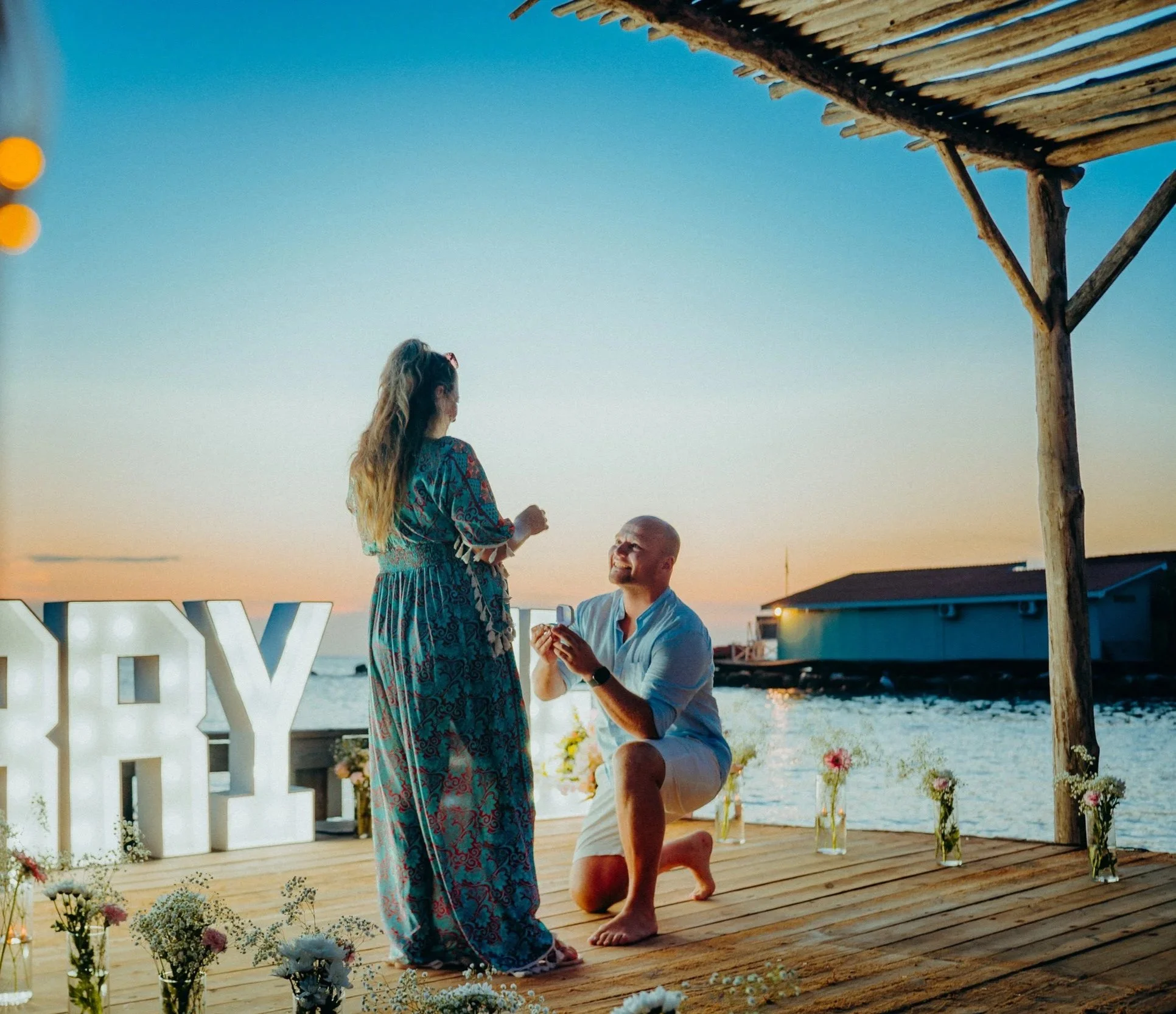 A woman in a colorful, patterned dress and sunglasses is proposing marriage to a man on a wooden deck by the water during sunset. The man is kneeling with a ring, smiling up at her. The scene is decorated with flowers and small vases, with a sign that reads 'LOVE' on the left, and a rustic hut or structure on the right.