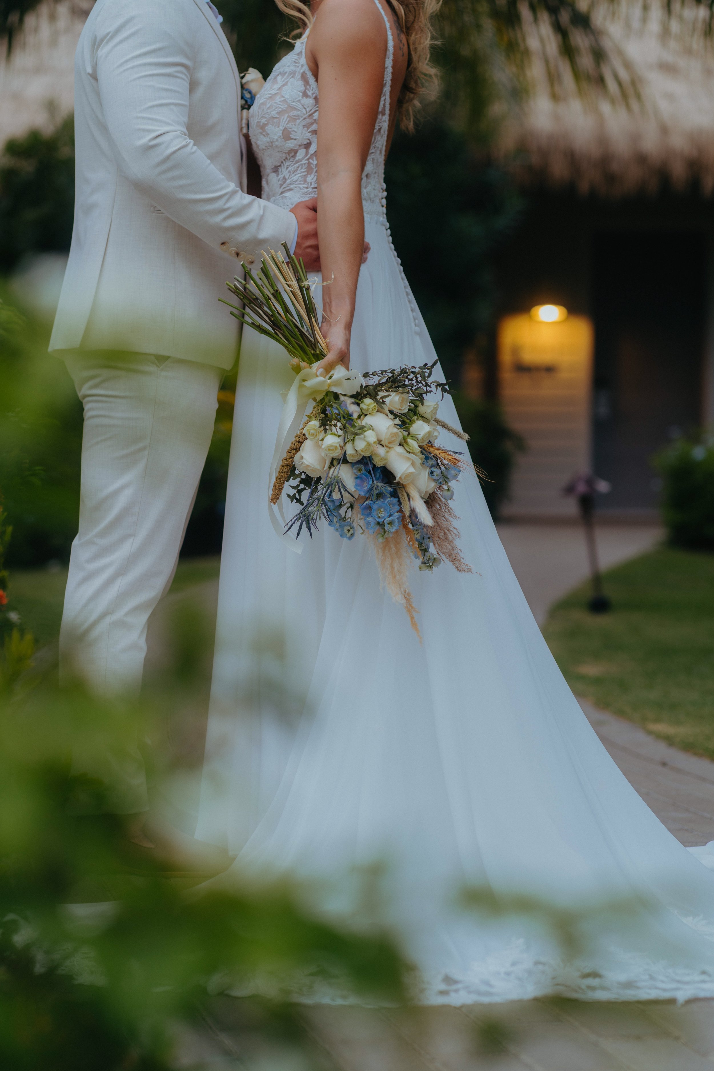 A bride and groom embrace outdoors, holding hands, with the bride holding a bouquet of white roses and greenery, as part of a wedding ceremony.