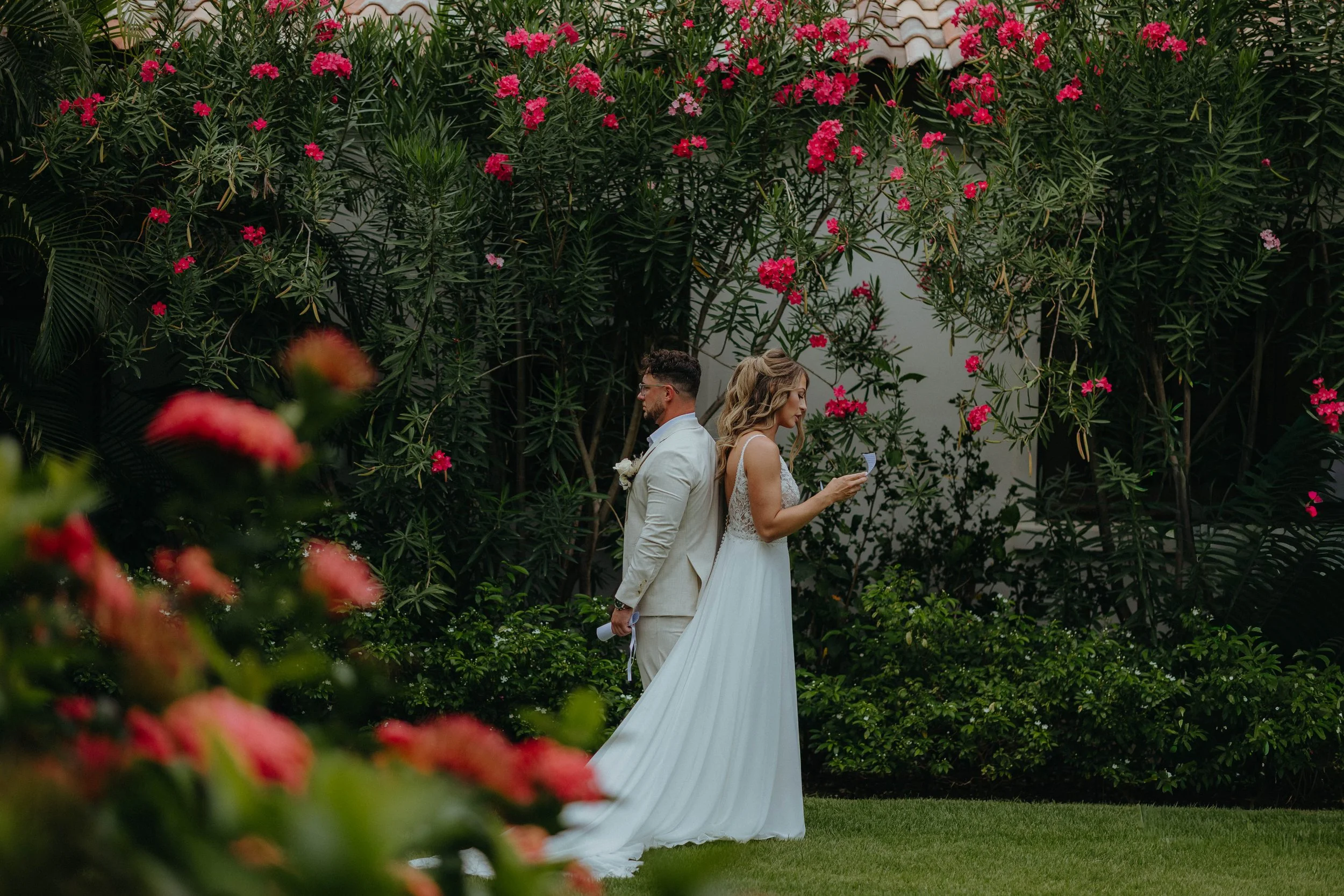 A bride and groom standing outdoors in front of large green plants with pink flowers, during a wedding ceremony.