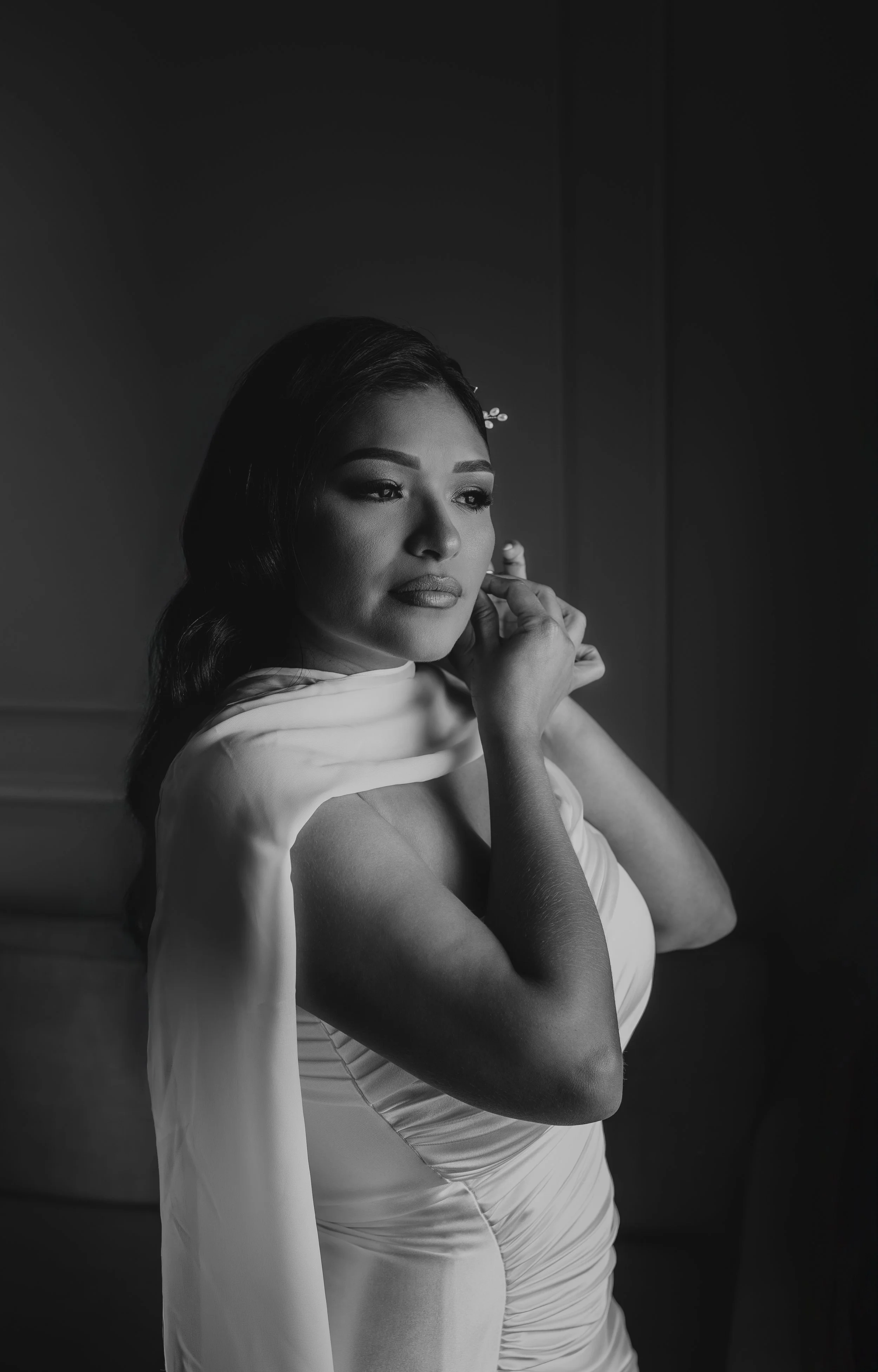 A black and white portrait of a woman with dark hair, makeup, and a confident expression, posing with her hand near her face and wearing a strapless dress.