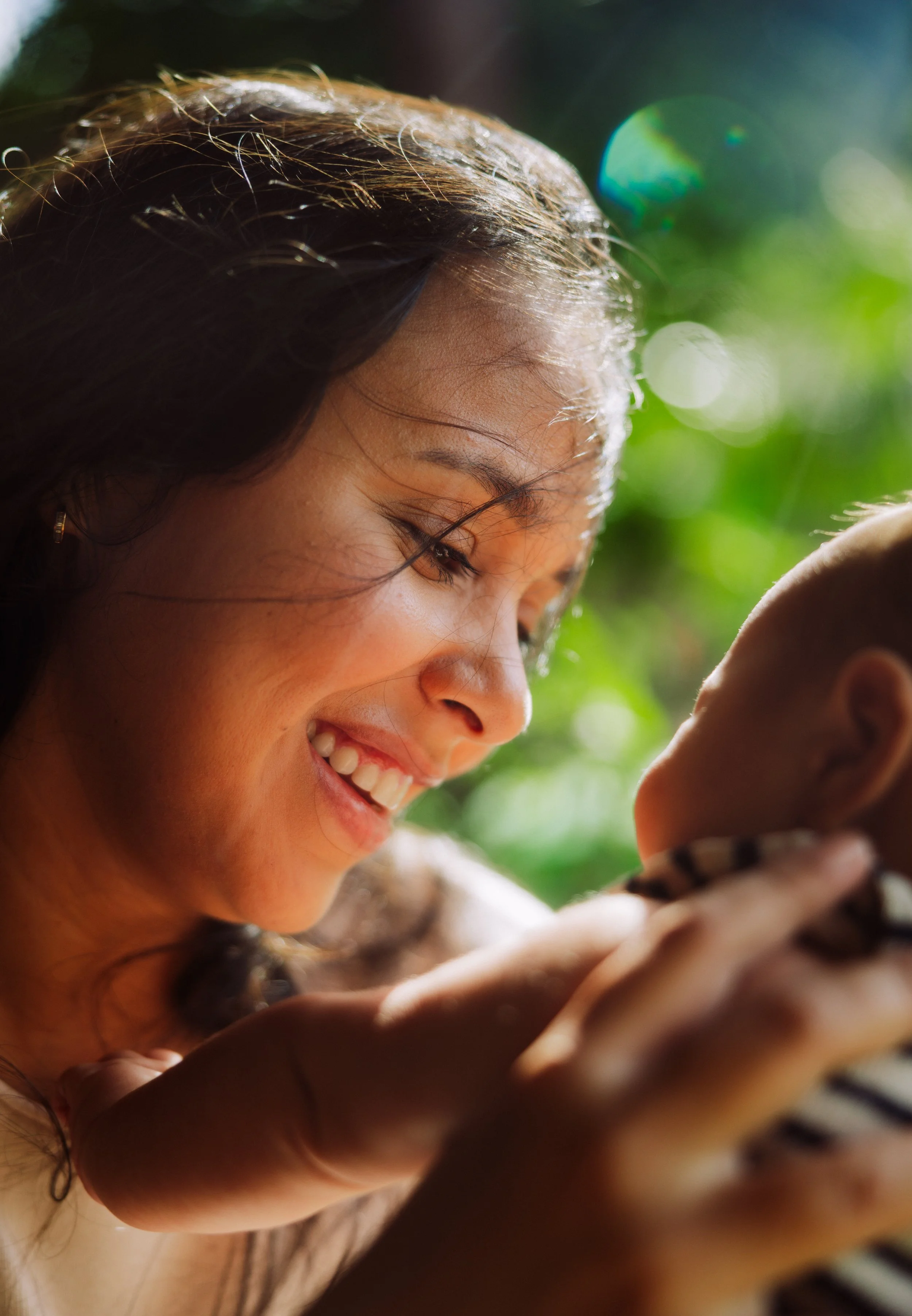A smiling woman with dark hair looking at a phone with a blurred child in the foreground, outdoors with greenery and sunlight.