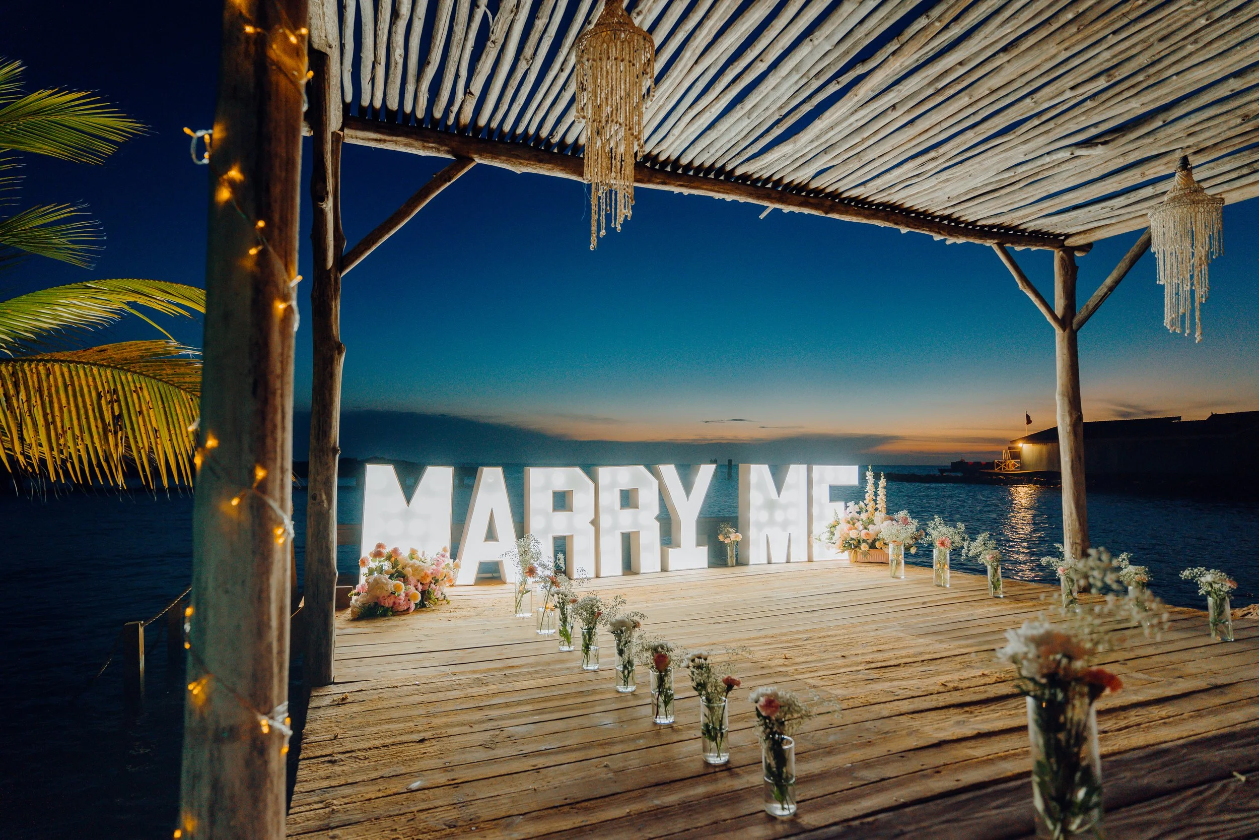 A decorated outdoor wedding setup on a wooden platform by a body of water at sunset, featuring large illuminated letters spelling 'MARRY ME', floral arrangements in vases, bamboo overhead structure, and hanging chandeliers with string decorations.