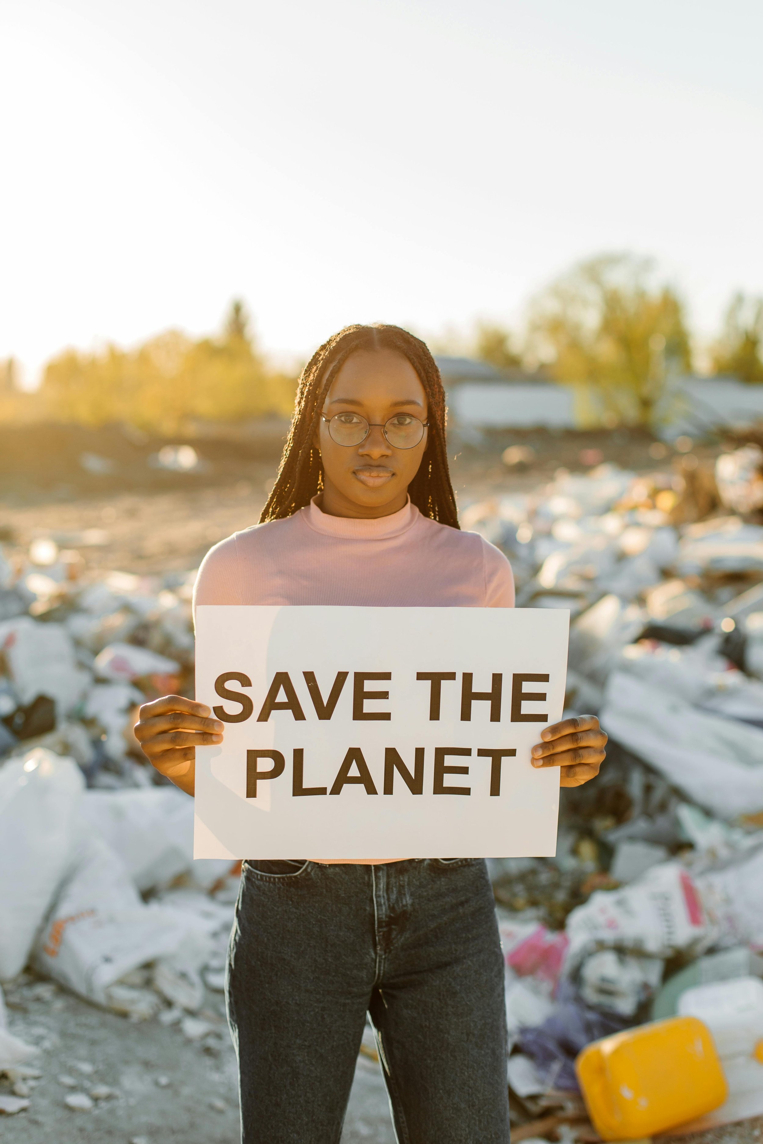 Young woman holding a sign that says 'Save the Planet' in a landfill or trash area at sunset.