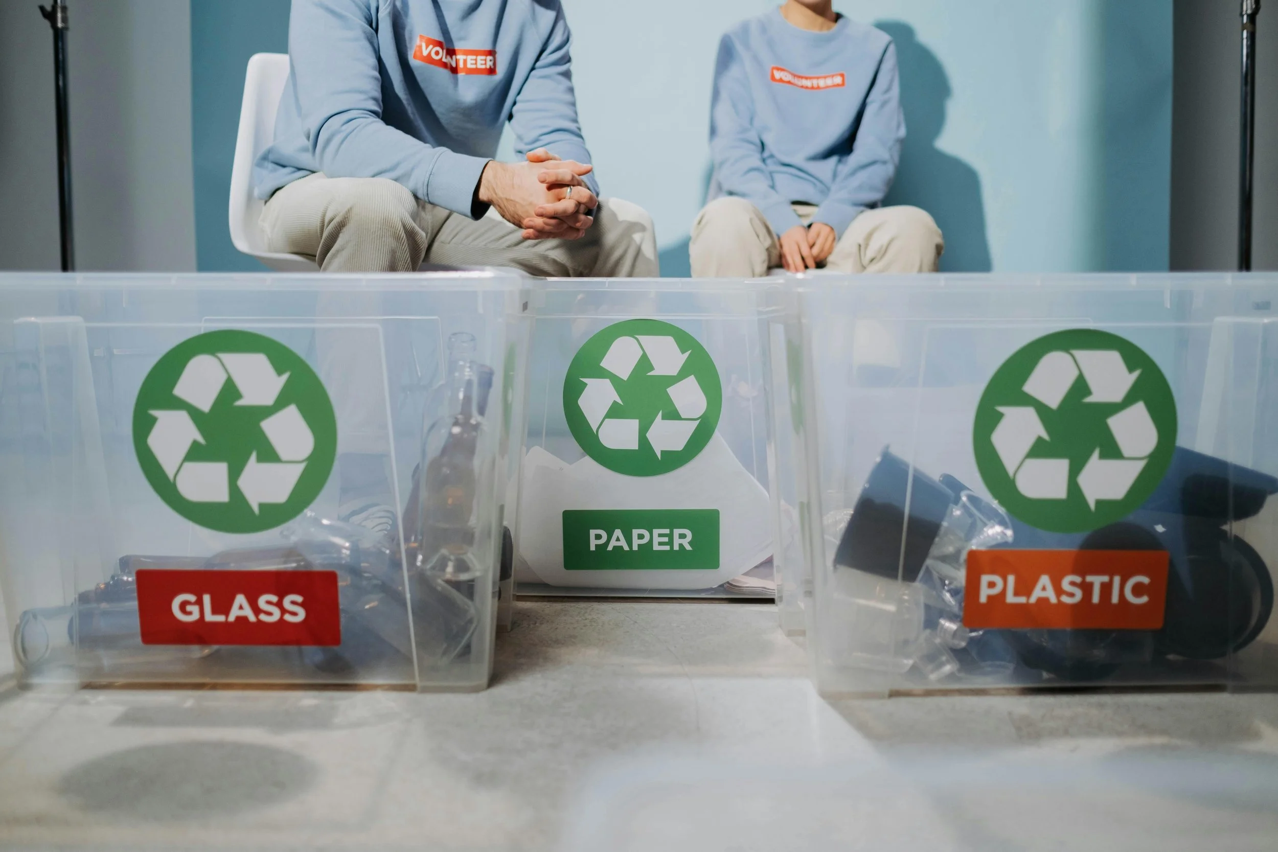 Two people wearing light blue volunteer shirts sitting behind clear recycling bins labeled for glass, paper, and plastic, with various recyclable items inside.