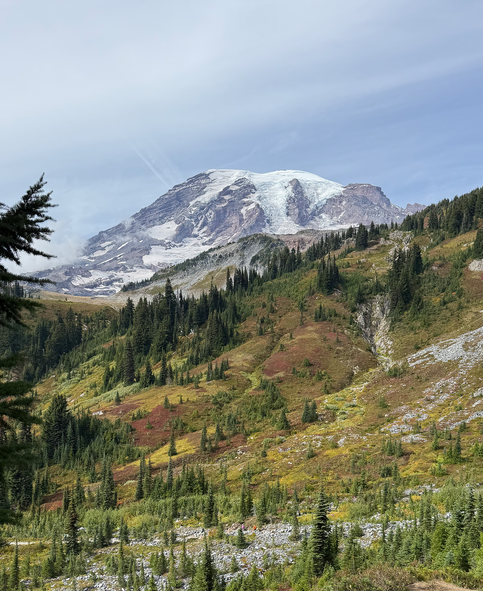 A snow-capped mountain in the distance with green forested hills in the foreground and a partly cloudy sky above.