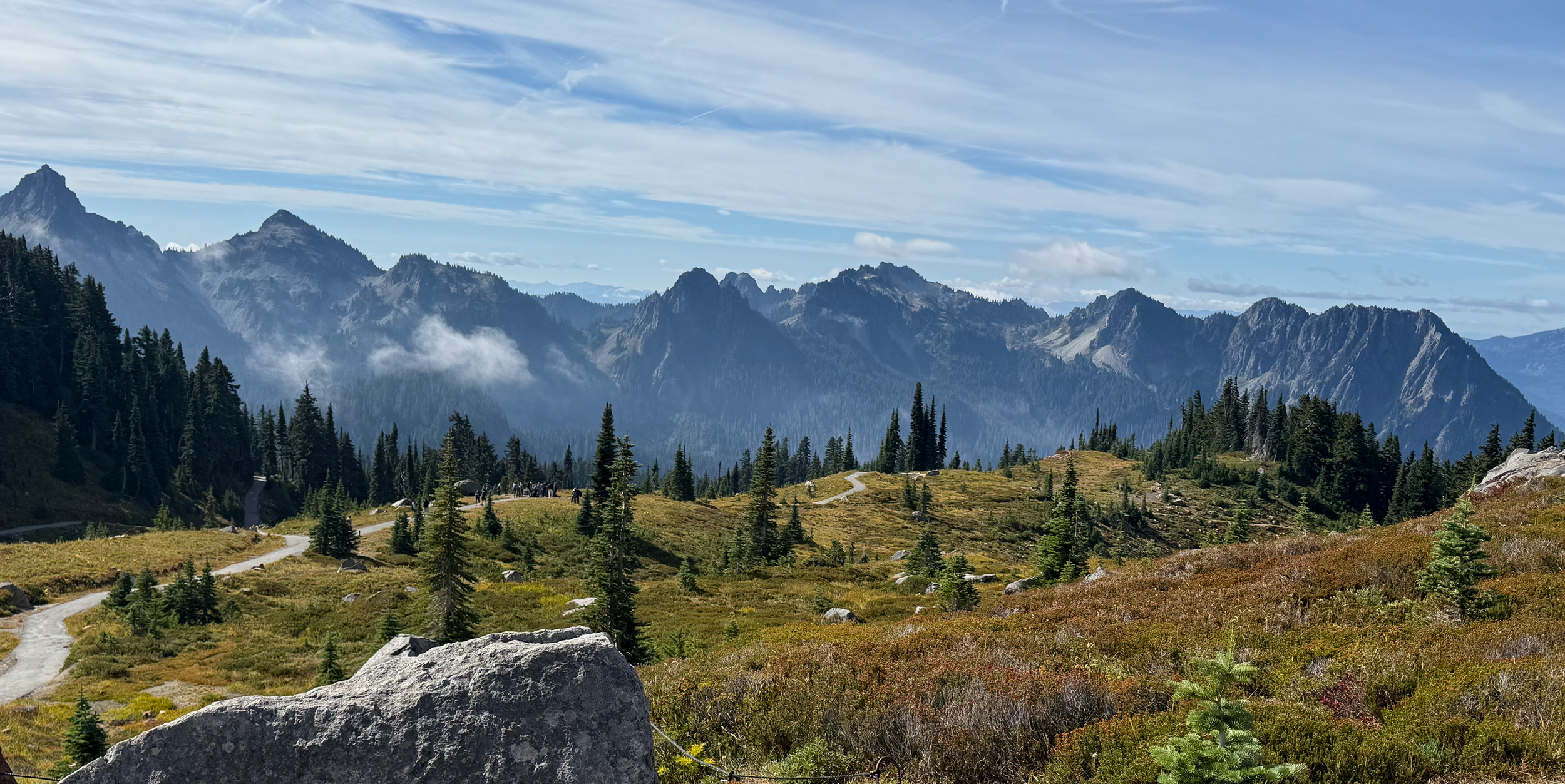A scenic mountain landscape featuring a range of jagged mountains, a foreground of dense evergreen trees, a grassy trail winding through the terrain, with partly cloudy skies and patches of mist or clouds around the mountains.