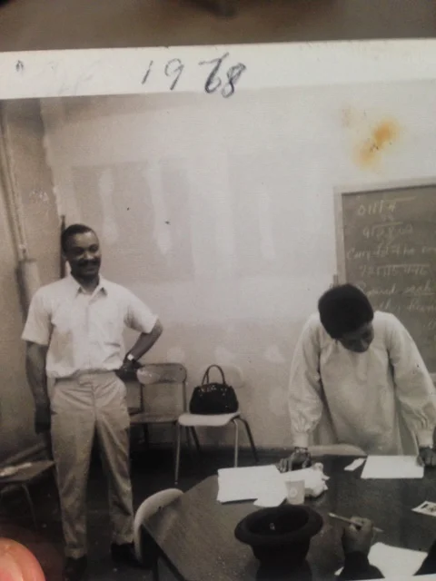 Brisa Builders Corp. Two men in a classroom, one standing with hands on hips and the other leaning over a table writing, with a blackboard and chairs in the background, dated 1968.