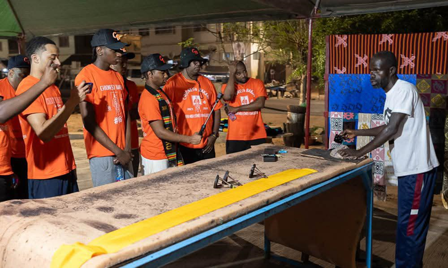 Brisa Builders Corp. A group of young men in orange shirts and hats listening to a man in a white shirt explaining something at an outdoor event at night.