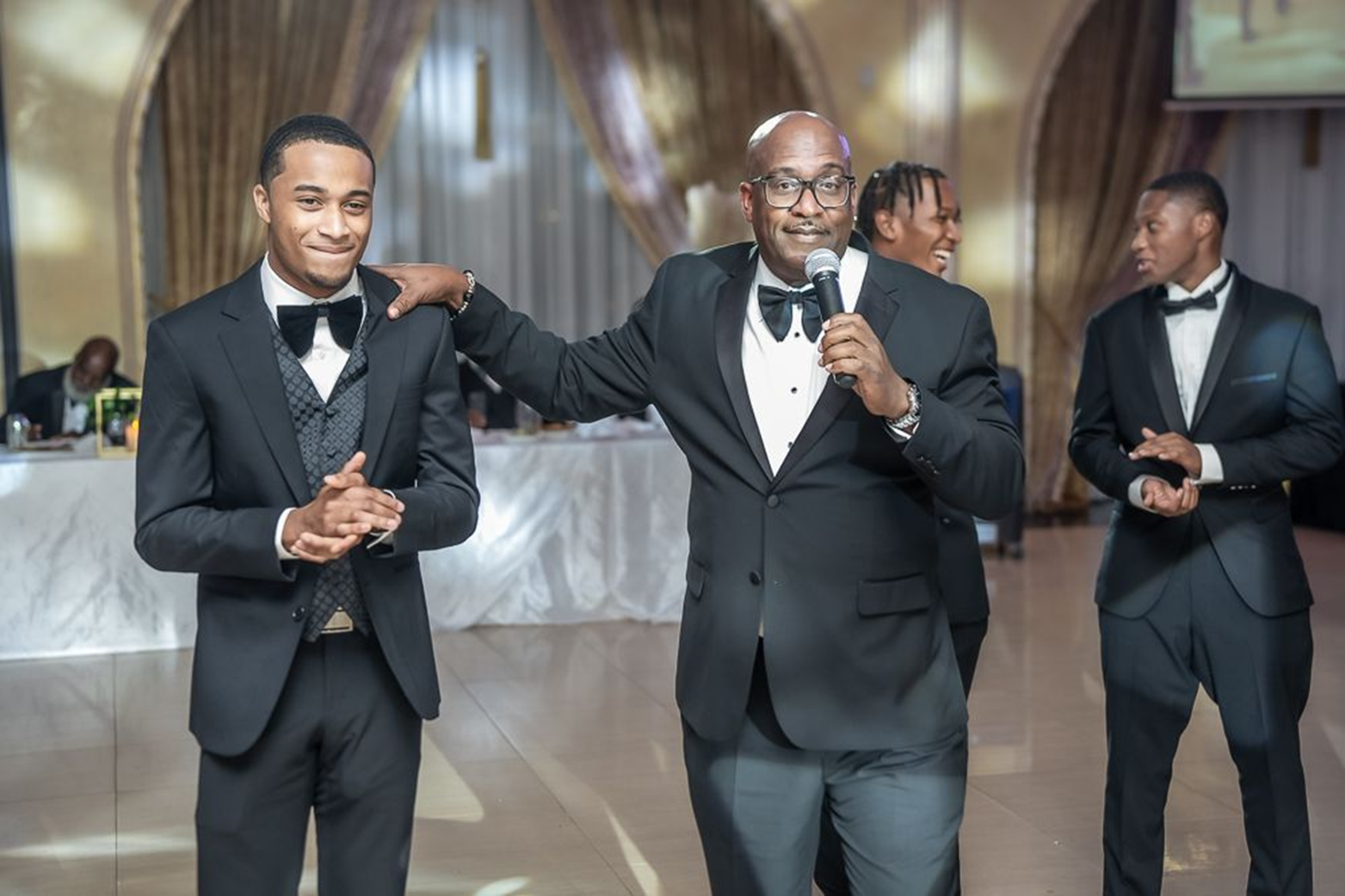 A man in a tuxedo speaking into a microphone with a young man in a tuxedo standing beside him during a celebration at a wedding reception.