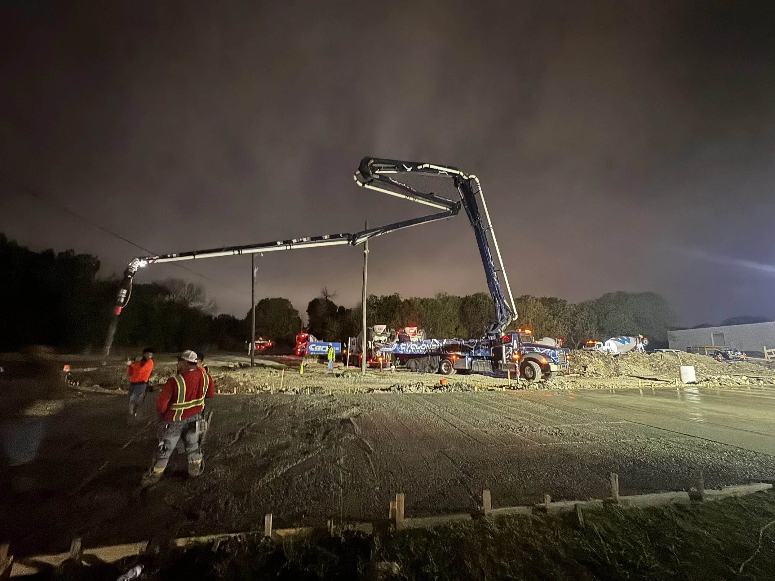 Nighttime construction scene showing a large concrete pump truck extending its arm above ground, with workers wearing safety vests and helmets at a construction site.