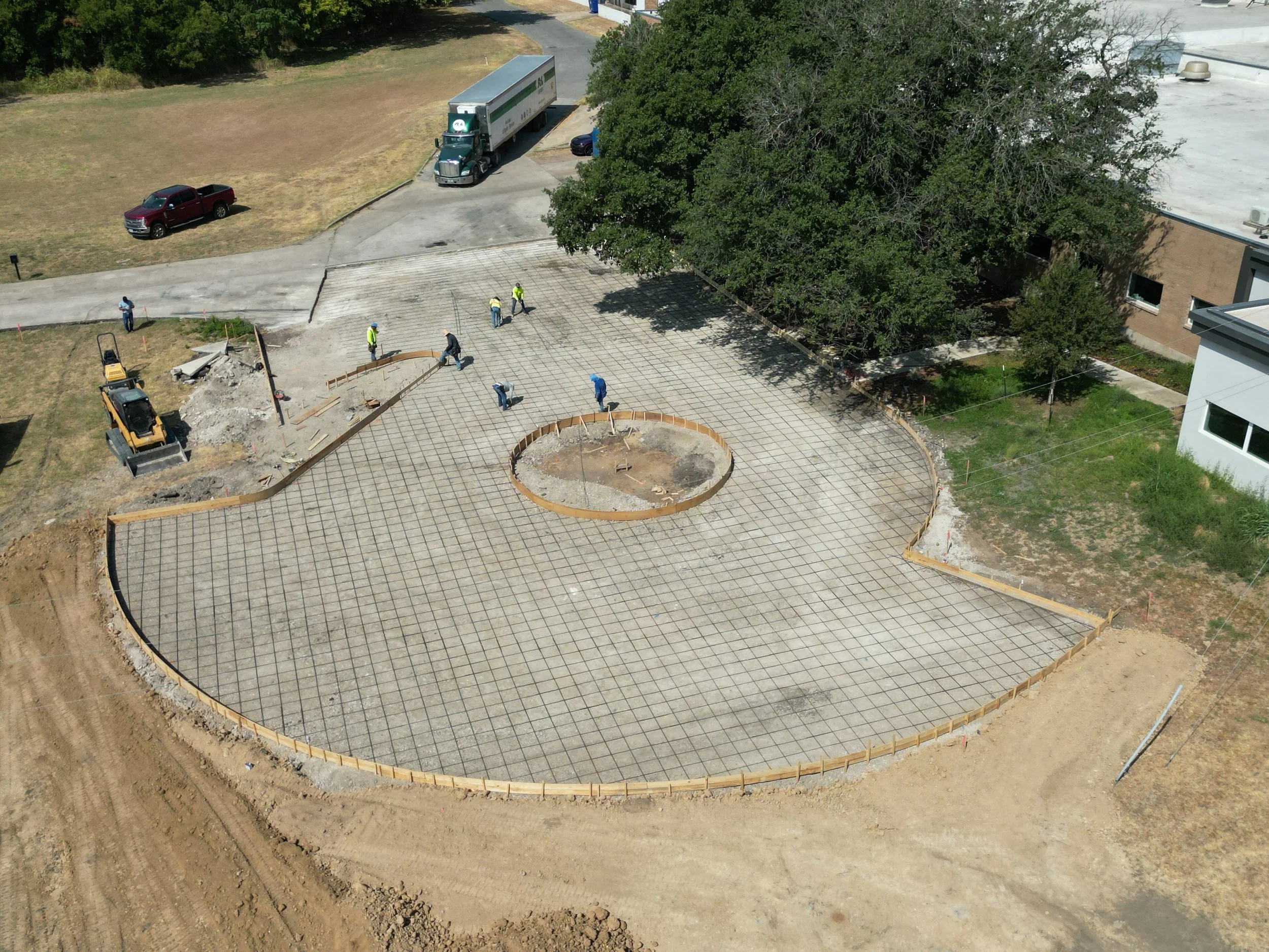 Construction workers are working on a concrete slab with a circular hole in the center, surrounded by a curved wooden border. There is construction equipment and dirt around the site, and a large tree is to the right of the construction area.