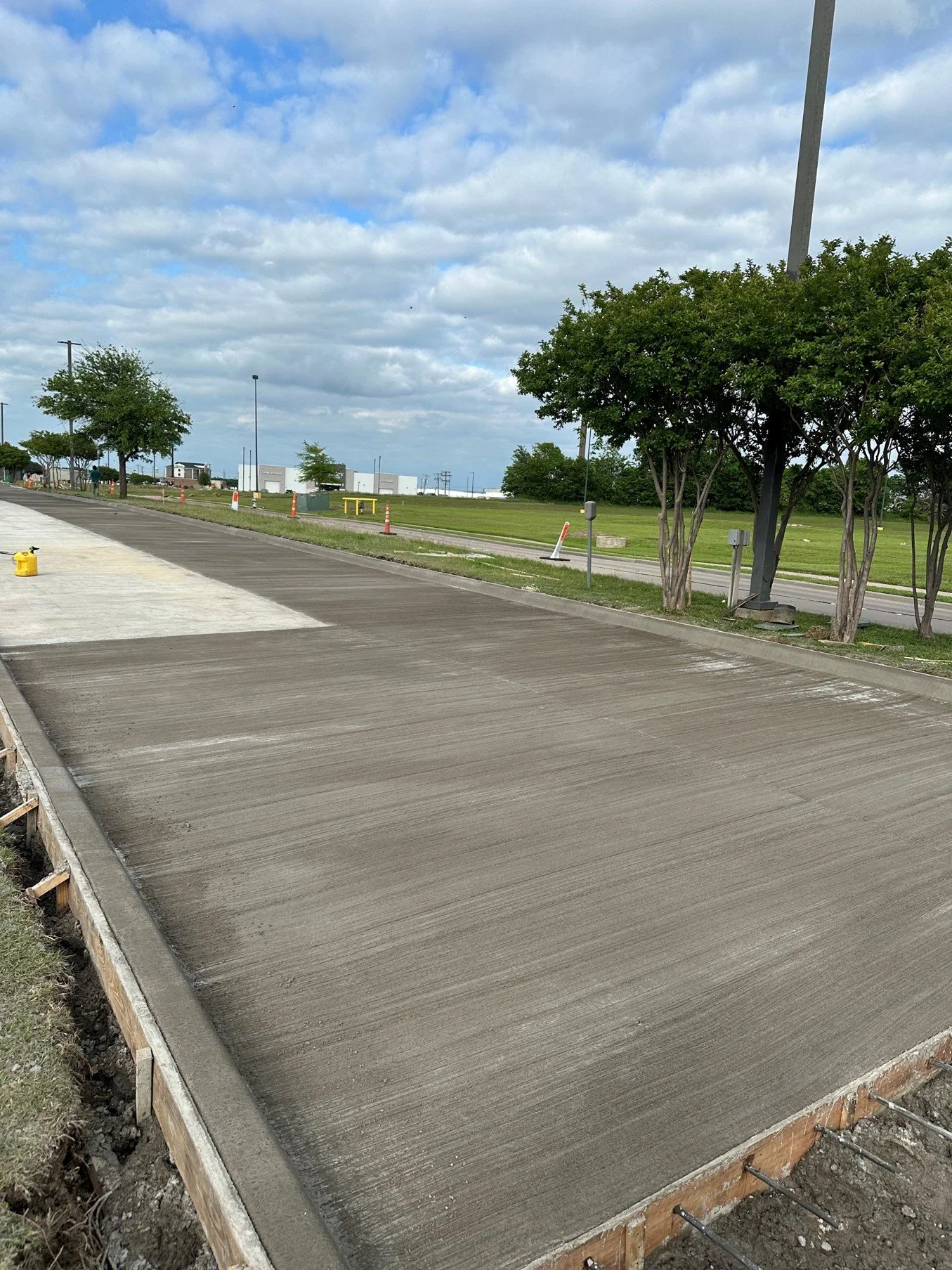 Freshly poured concrete sidewalk under a partly cloudy sky with trees and construction cones in the background.